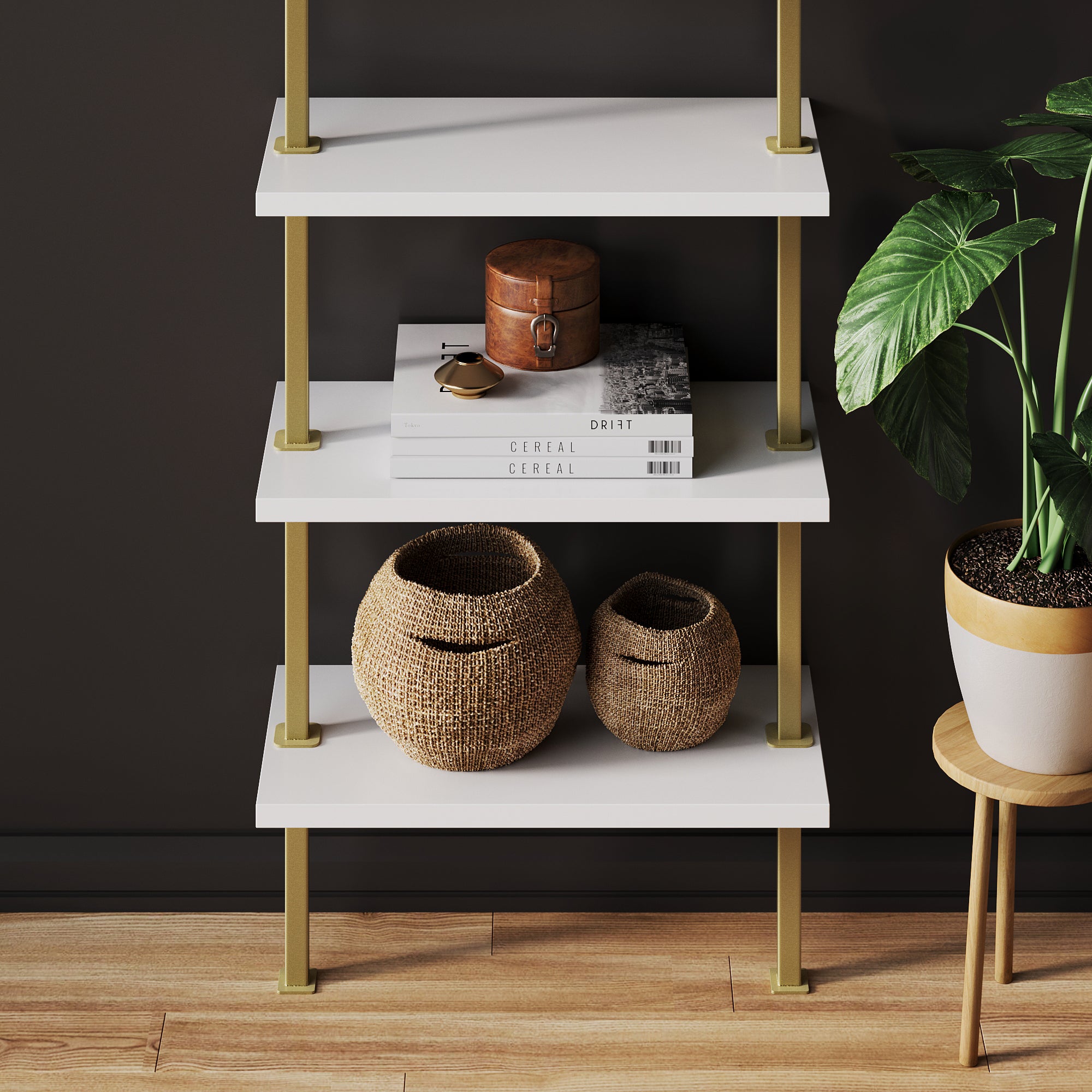 A white three-tier shelf with gold supports holds books, a small wooden box, two woven baskets, and is next to a green potted plant on a wooden floor against a dark wall.