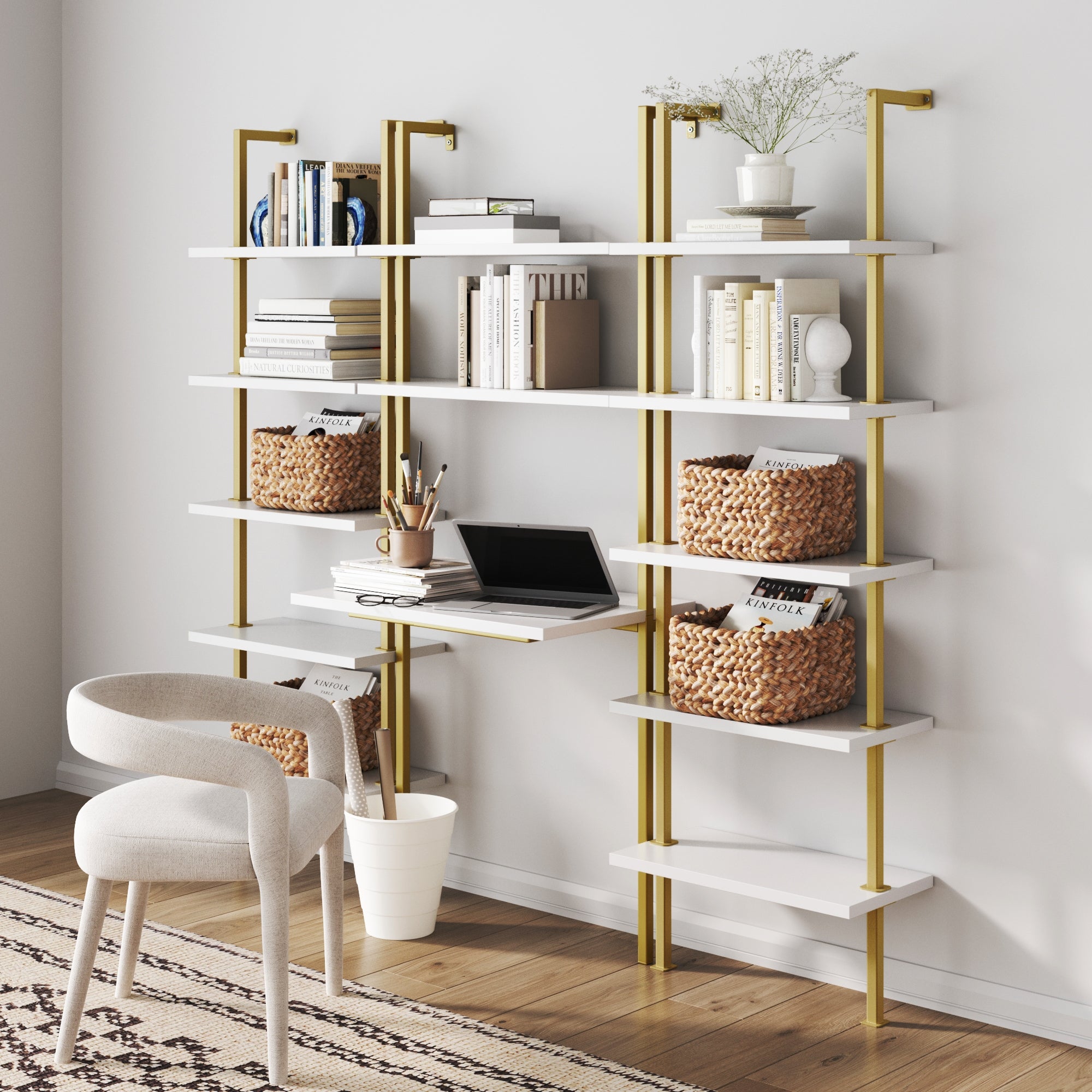 The Nathan James Glam Wall Mount Metal Desk and Bookcase Set features gold frames and white shelves, stylishly displaying books, baskets, plants, and decor against a white wall. A white chair and patterned rug sit on the wooden floor in front.