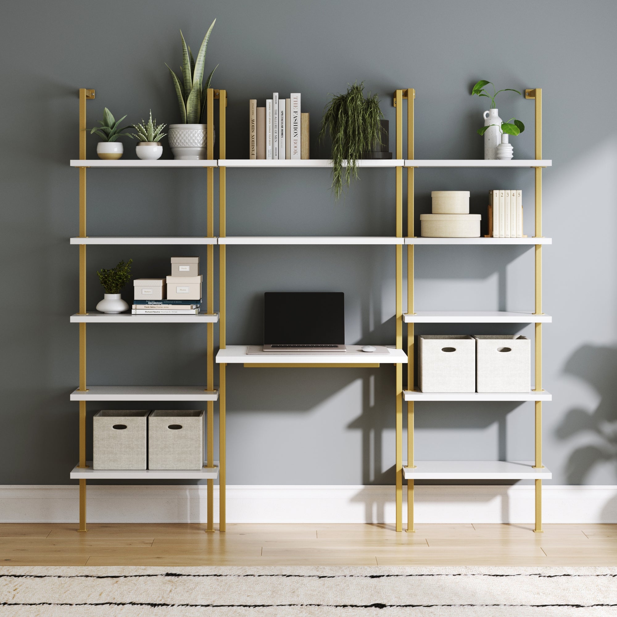 A modern workspace showcases the Nathan James Glam Wall Mount Metal Desk and Bookcase Set with gold supports, displaying books, plants, boxes, and baskets. A laptop rests on the built-in desk against a gray wall above a light wood floor and beige rug.