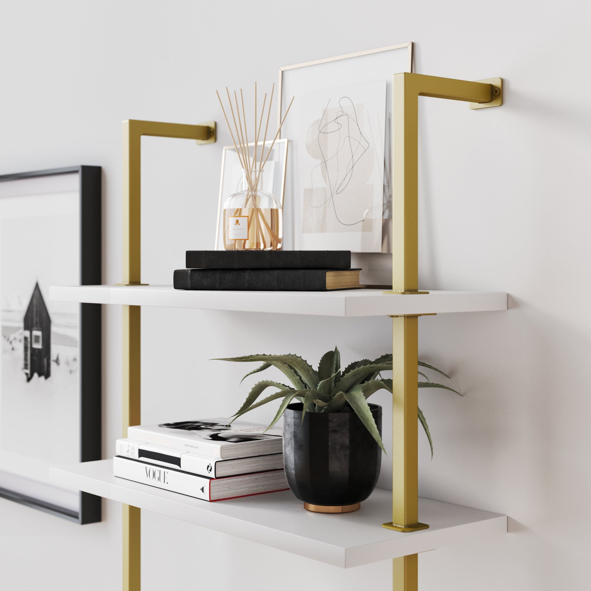 A white wall shelf with gold brackets holds stacked books, a black potted plant, a photo frame, a reed diffuser, and abstract art. Another black-and-white photo is mounted on the wall beside the shelf.