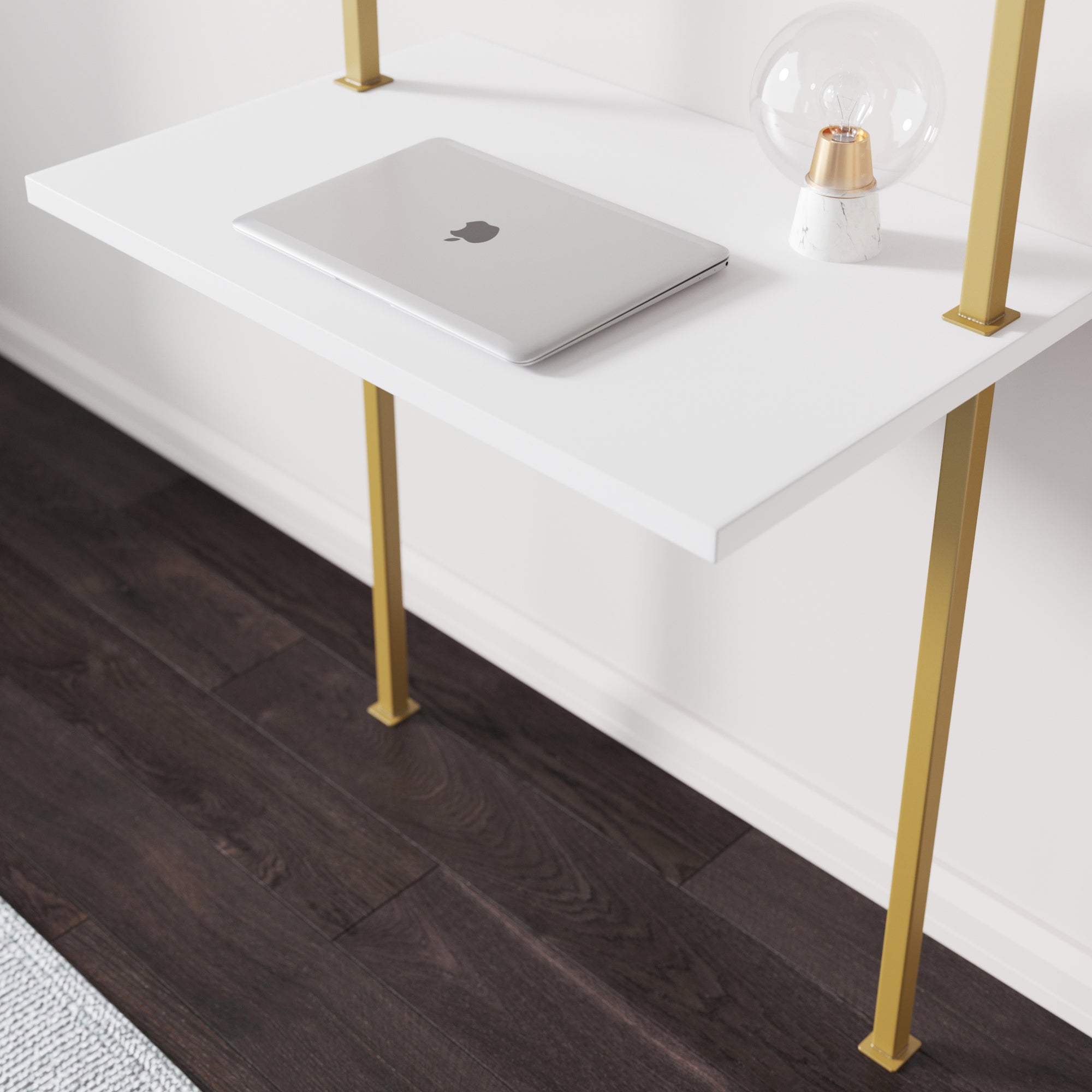 A closed silver MacBook sits on a white desk with gold legs, next to a modern clear-glass table lamp, on a dark wood floor.