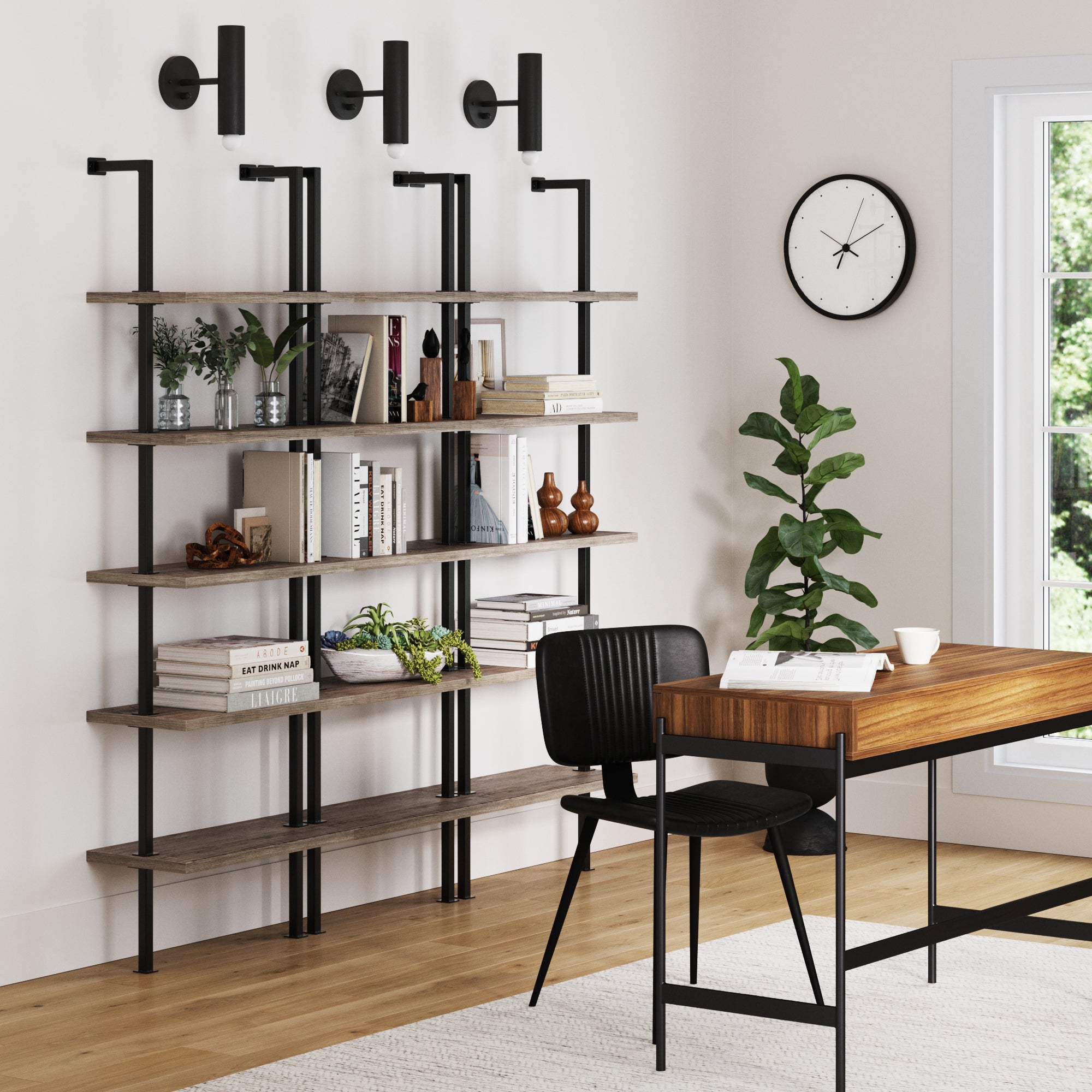A modern home office features a wooden desk, black chair, wall clock, potted plant, and a set of three Nathan James 5-Shelf Ladder Bookshelves in Gray Oak displaying books and decor against a light wall with three black sconces.