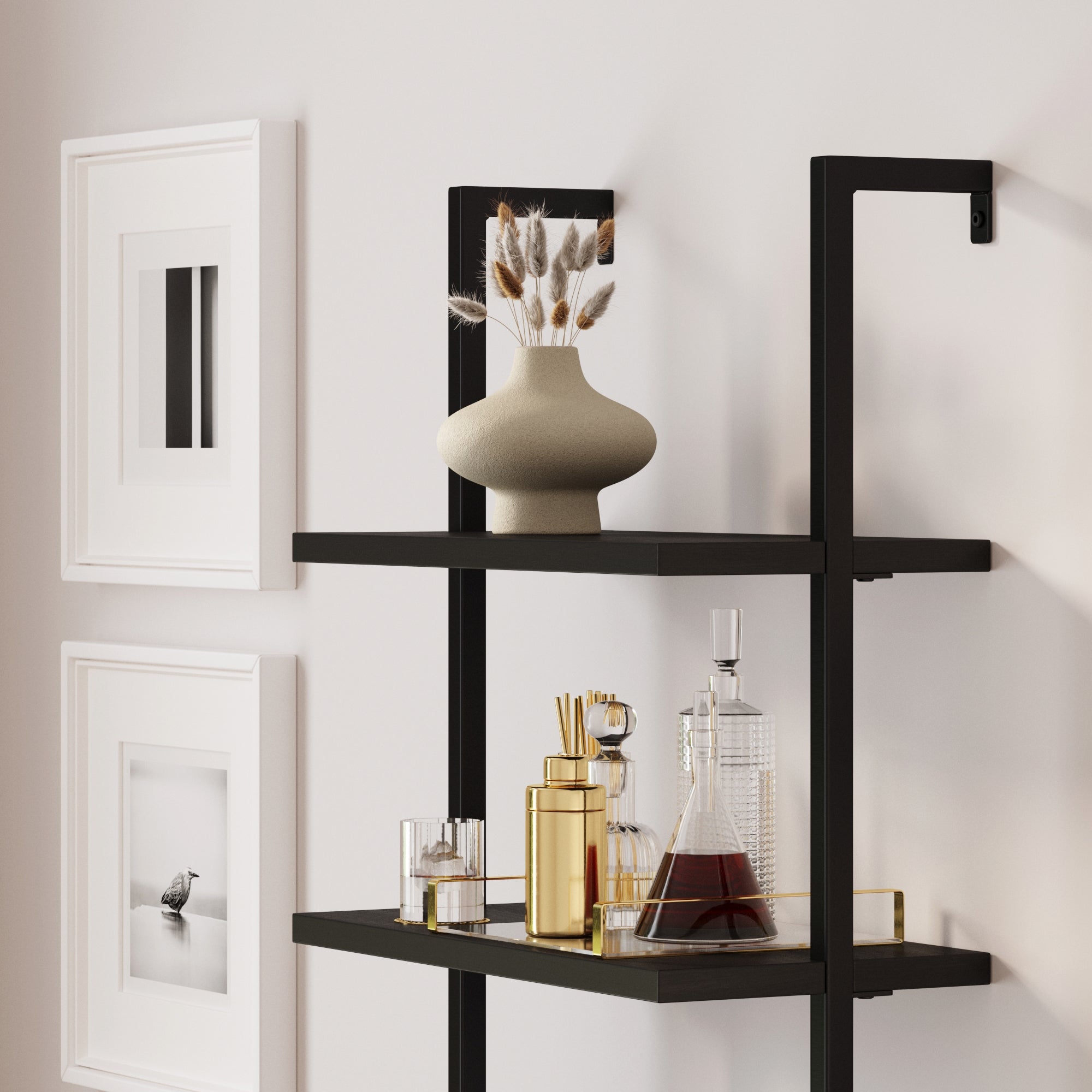 A modern black wall-mounted shelf displays decorative glass bottles, a decanter with dark liquid, a gold tray, and a beige vase with dried flowers. Minimalist framed art hangs on the white wall nearby.
