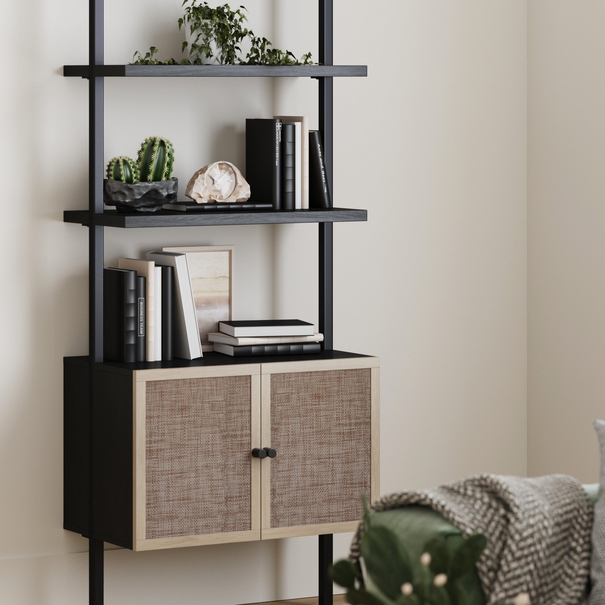 A modern black shelving unit with books, plants, and decorative objects, above a cabinet with woven doors. A cozy blanket and green plant are partially visible in the foreground.