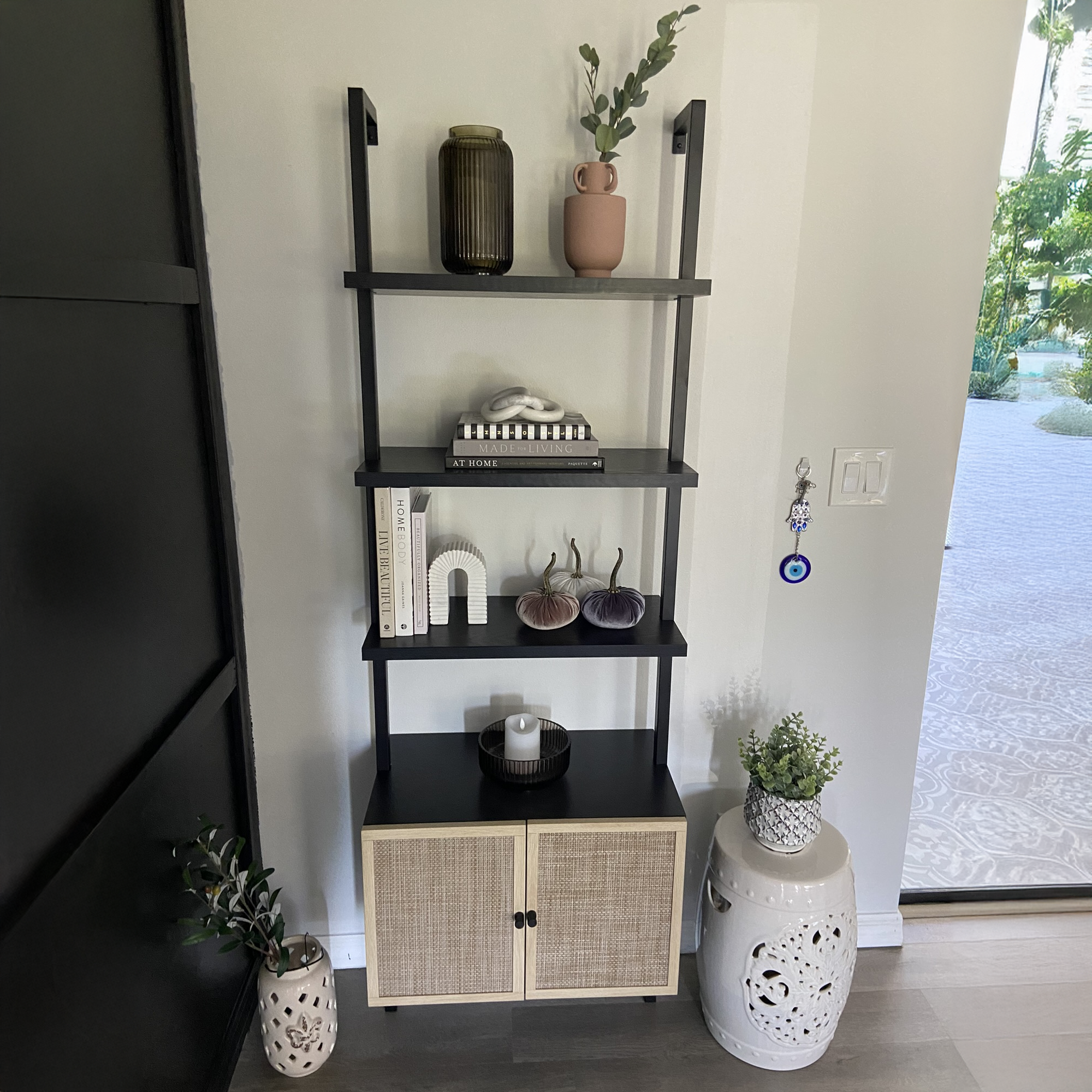 A black shelving unit with books, vases, art objects, and plants stands against a white wall. A white ceramic stool and potted plant are to the right, near a door with a key holder and an evil eye charm.