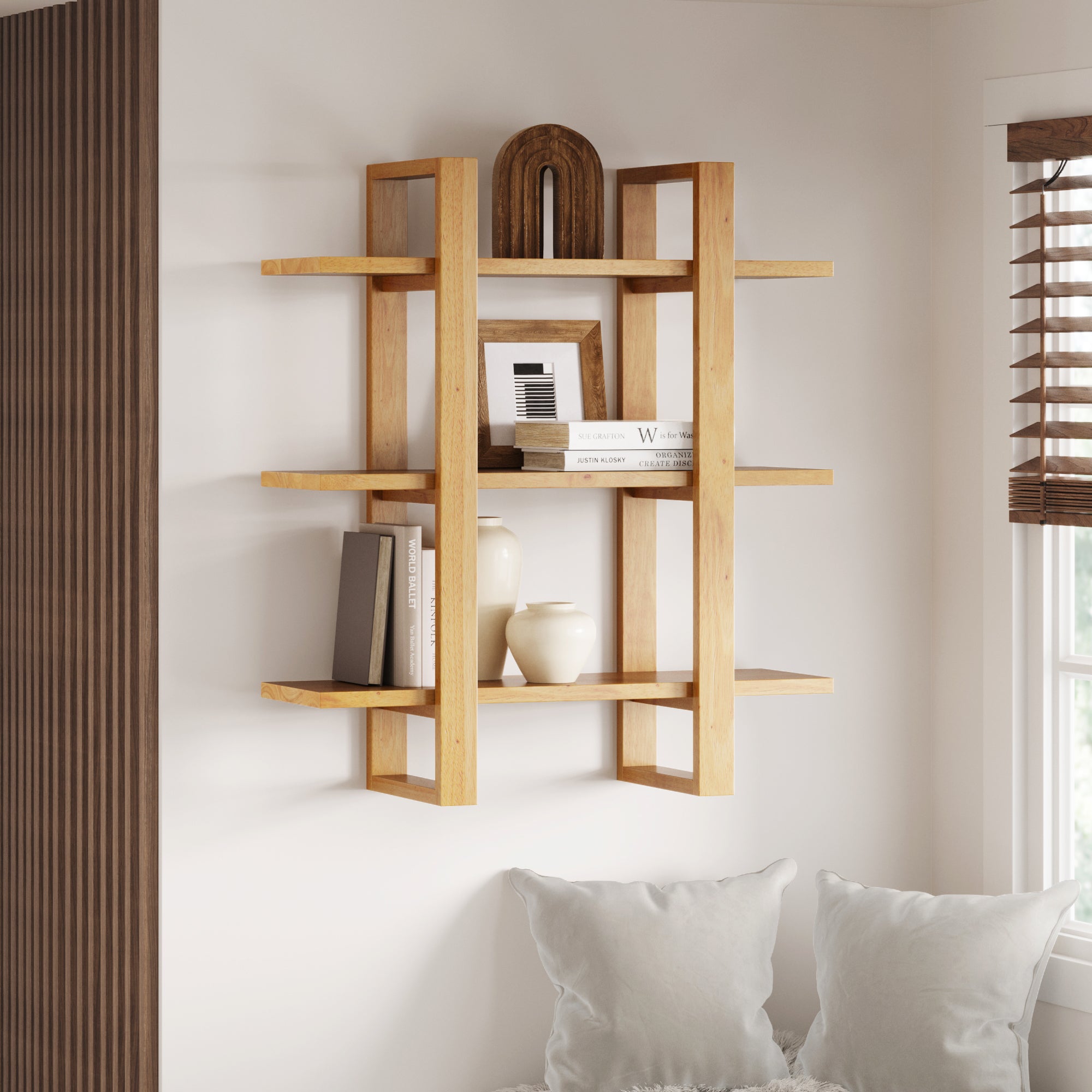 A modern wooden wall shelf with geometric design holds books, decorative vases, and an abstract sculpture. Below the shelf are white cushions, and sunlight streams through blinds on a nearby window.