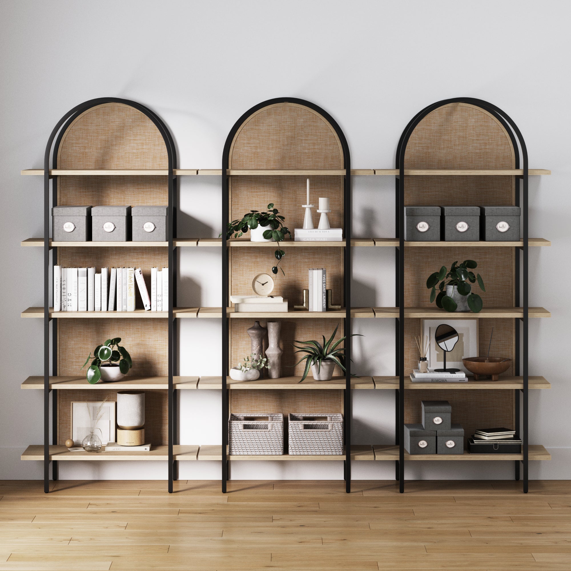 The Nathan James Boho Rattan & Wood Arched Bookshelves (Set of 3) display books, plants, baskets, decor, and art against a white wall on a wooden floor.