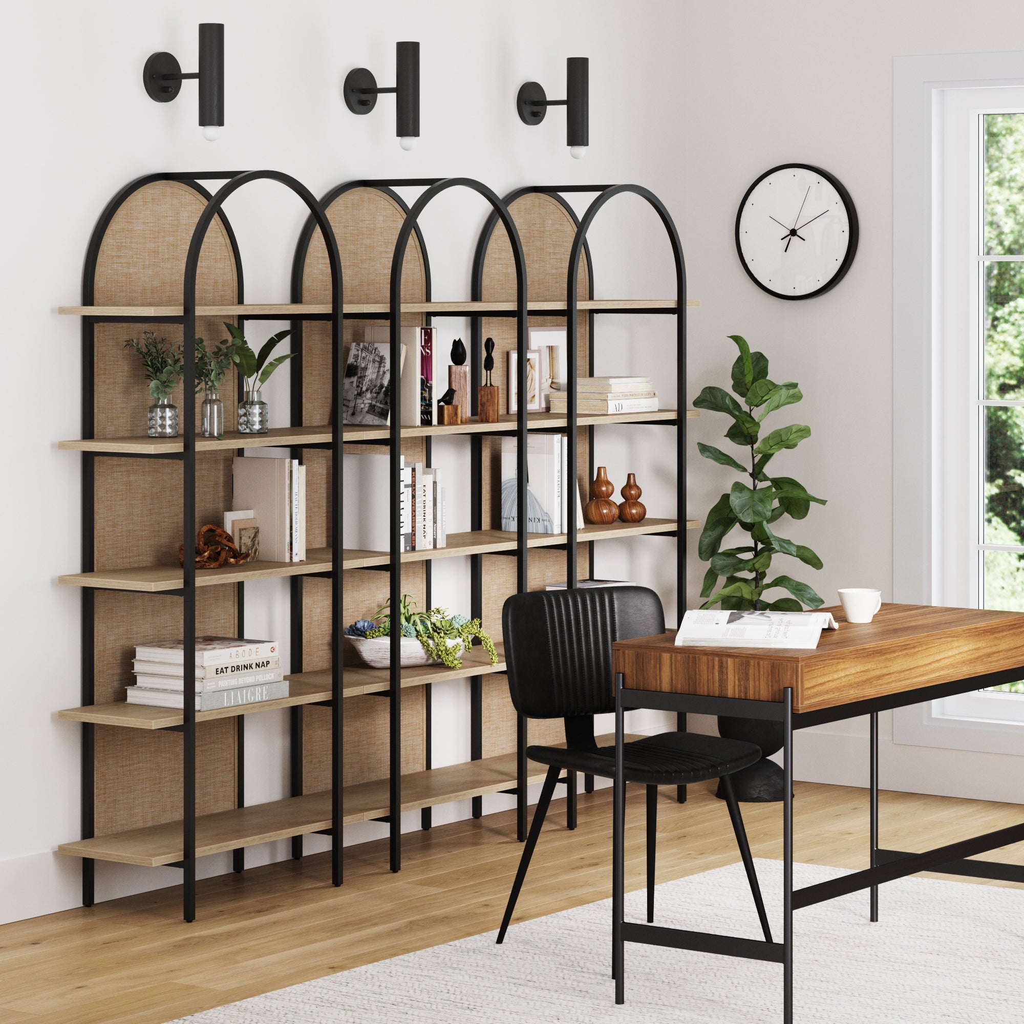 A modern home office with a wooden desk, black chair, Nathan James Boho Rattan & Wood Arched Bookshelves (Set of 3), books, plants, and decor. A wall clock, three black wall lights, and a large window complete the bright minimalist space.