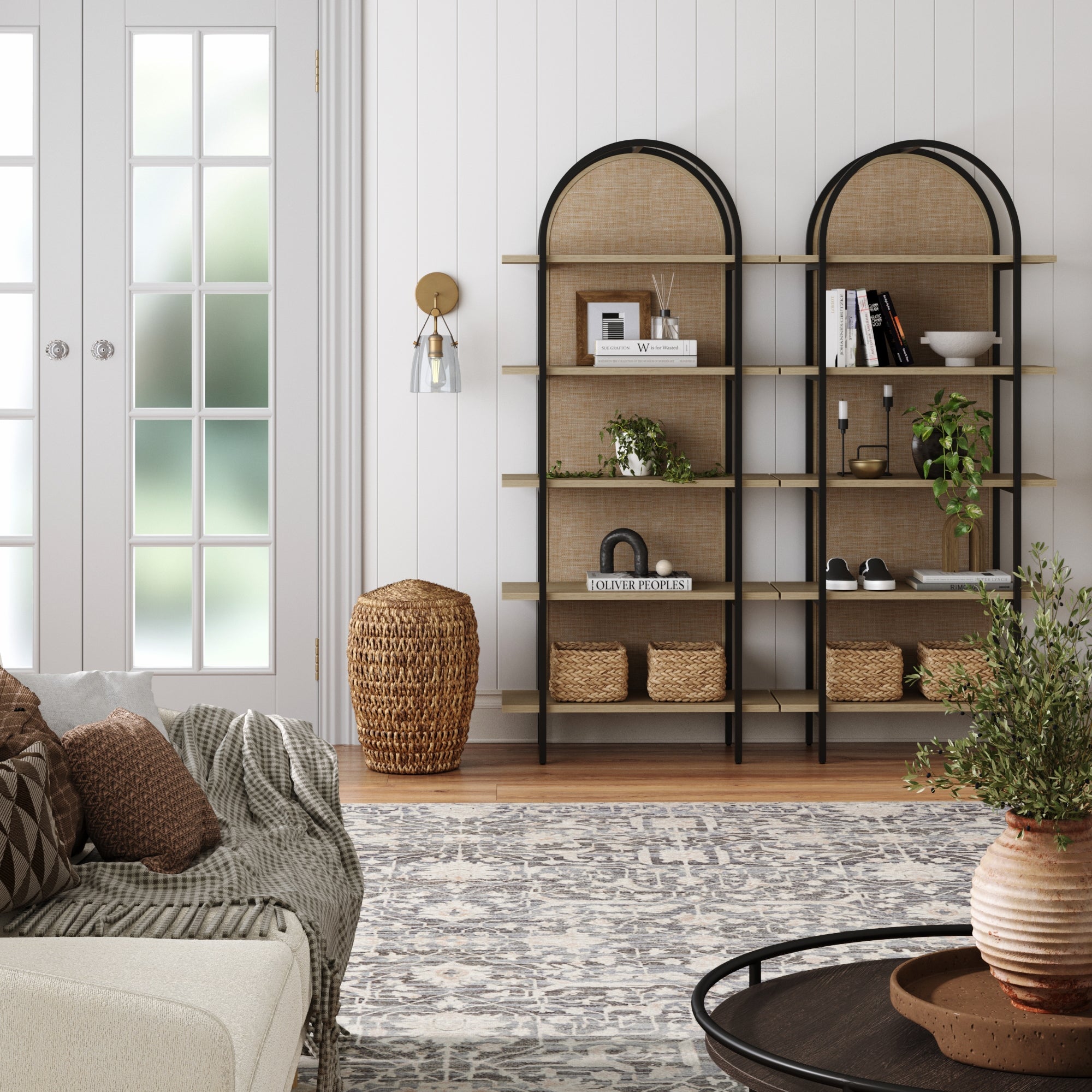 A cozy living room with a patterned rug, round coffee table, modern white sofa, and arched bookshelves decorated with books, baskets, and plants. Sunlight streams through glass double doors.