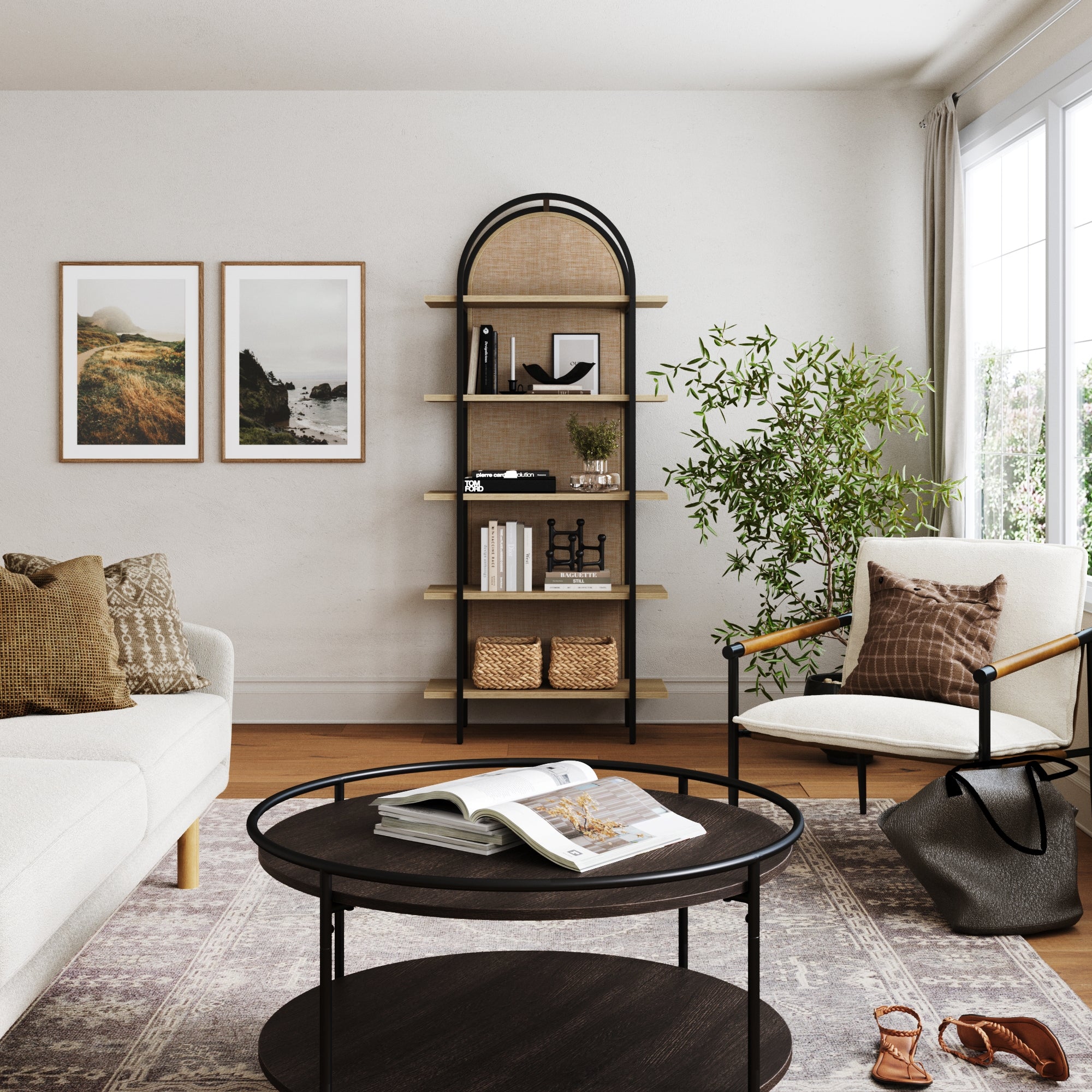 A modern living room features a white sofa, an armchair, a round black coffee table, and the Nathan James Boho Rattan & Wood Arched Bookshelf filled with decor and baskets, plus framed art, a large window, and a green plant in the corner.