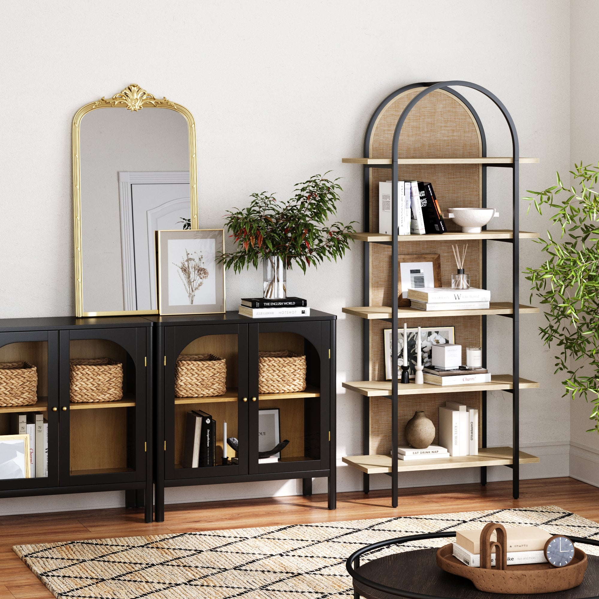 A stylish living room corner with black cabinets, a gold-framed mirror, a tall arched shelving unit with decor items, a potted plant, books, baskets, and a patterned rug on a wooden floor.