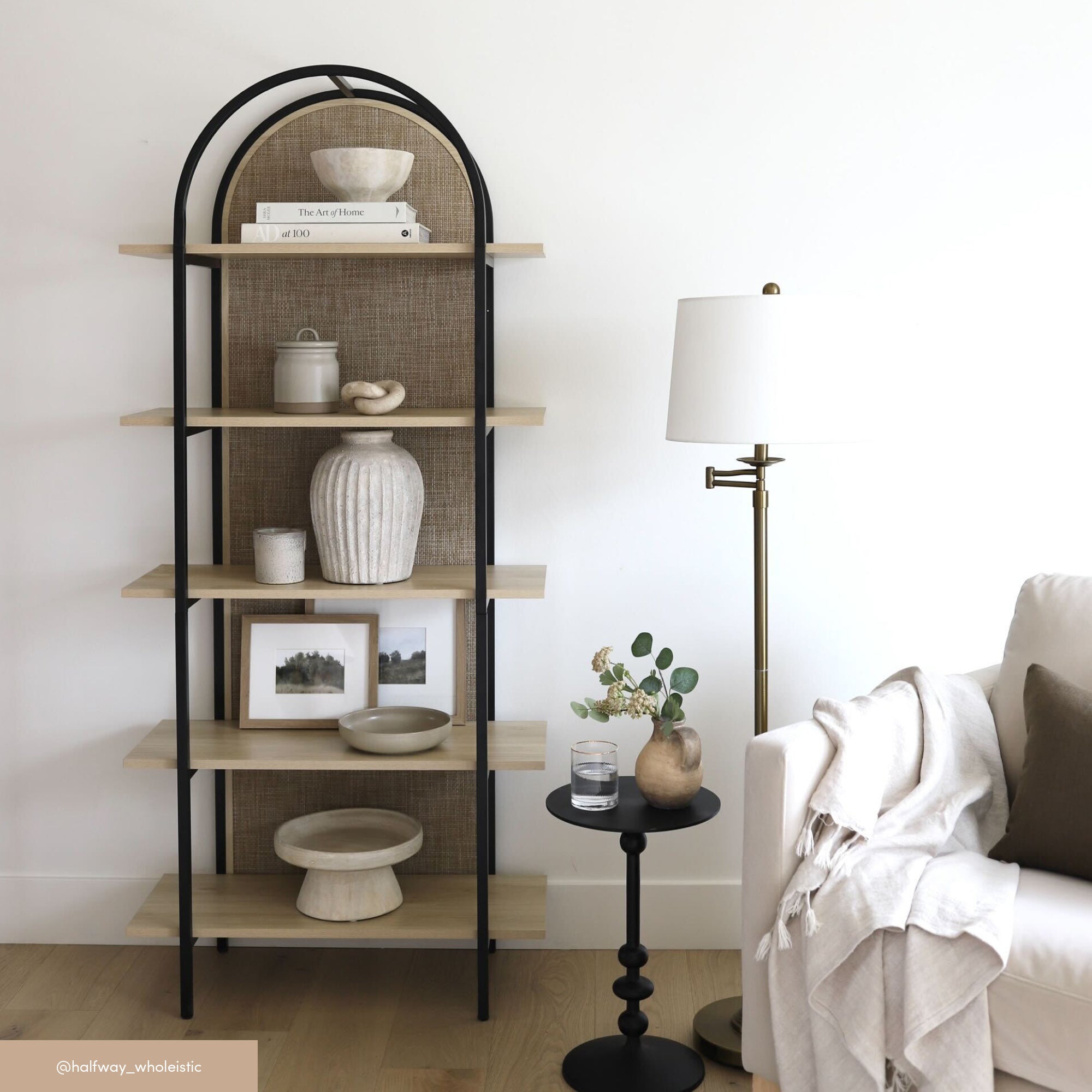 A modern living room corner with a black metal and wood open bookshelf holding vases, bowls, books, and framed art. Nearby, a brass floor lamp stands beside a white sofa draped with a beige throw and a small black round side table.