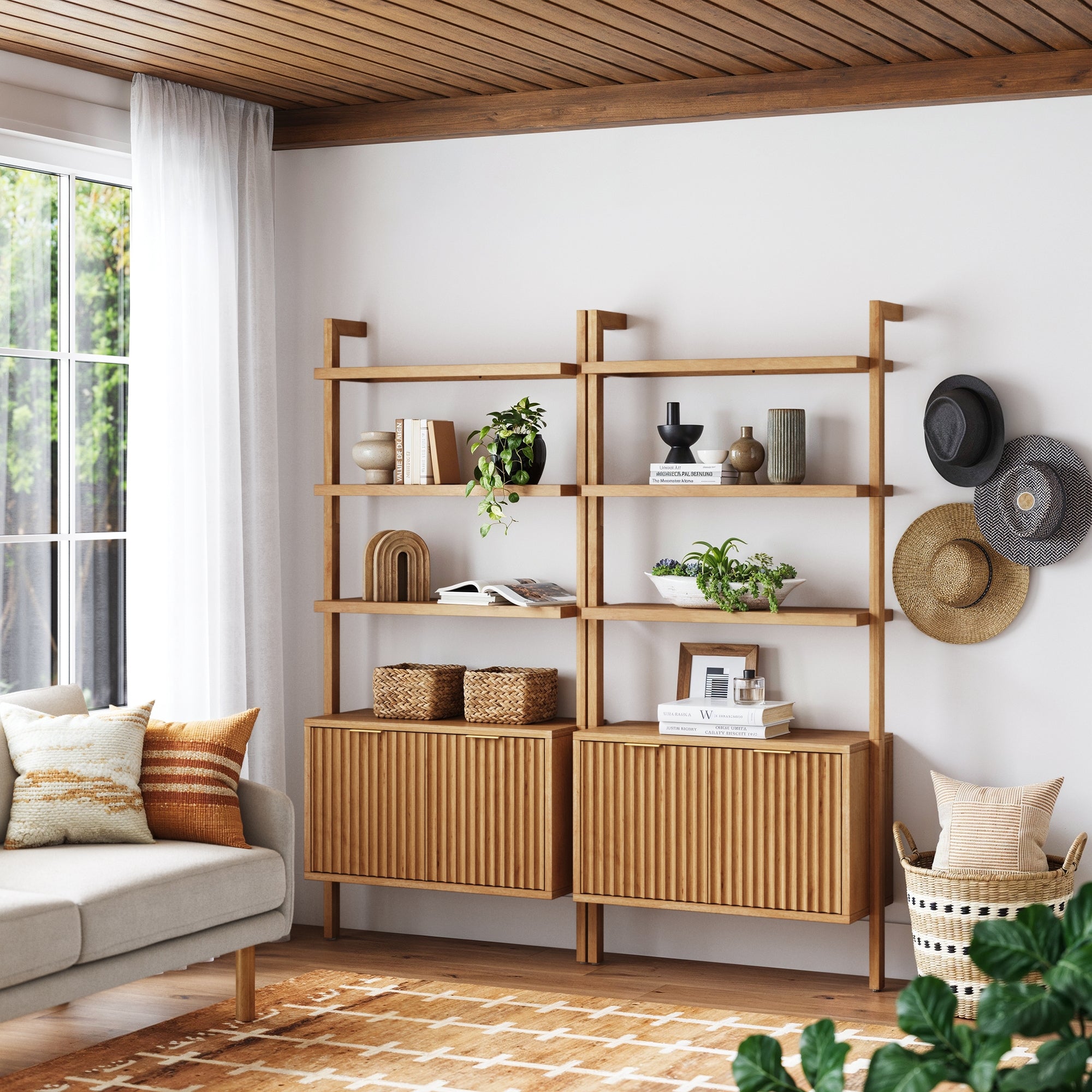 A modern living room features Nathan James Mid-Century Fluted Wall Bookshelves with Doors in Warm Pine (Set of 2) holding plants and decor, a beige sofa with patterned pillows, wall-mounted hats, white curtains, and a sunlit window.