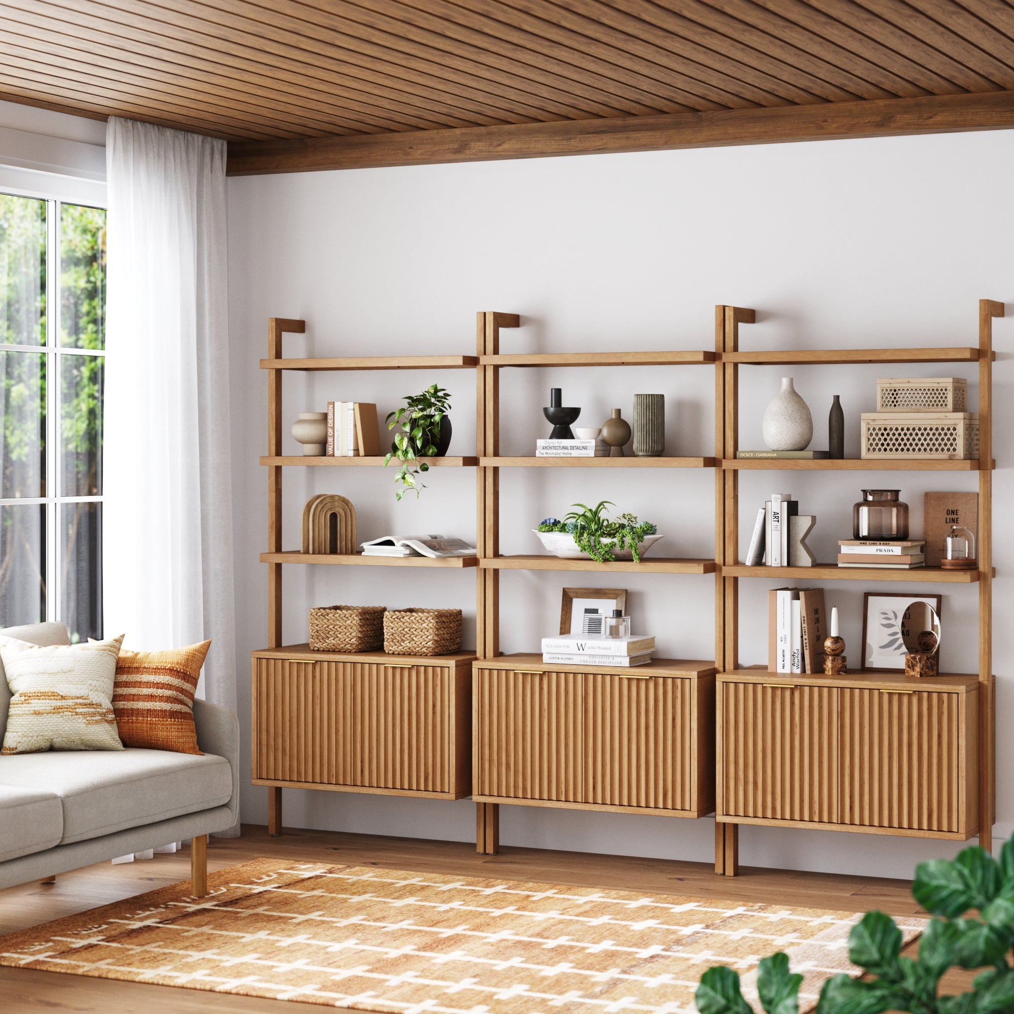 A modern living room features Nathan James Mid-Century Fluted Wall Bookshelves with Doors in Warm Pine (Set of 3), showcasing decor, books, and plants. A light sofa with pillows, a patterned rug, and large windows complete the space.