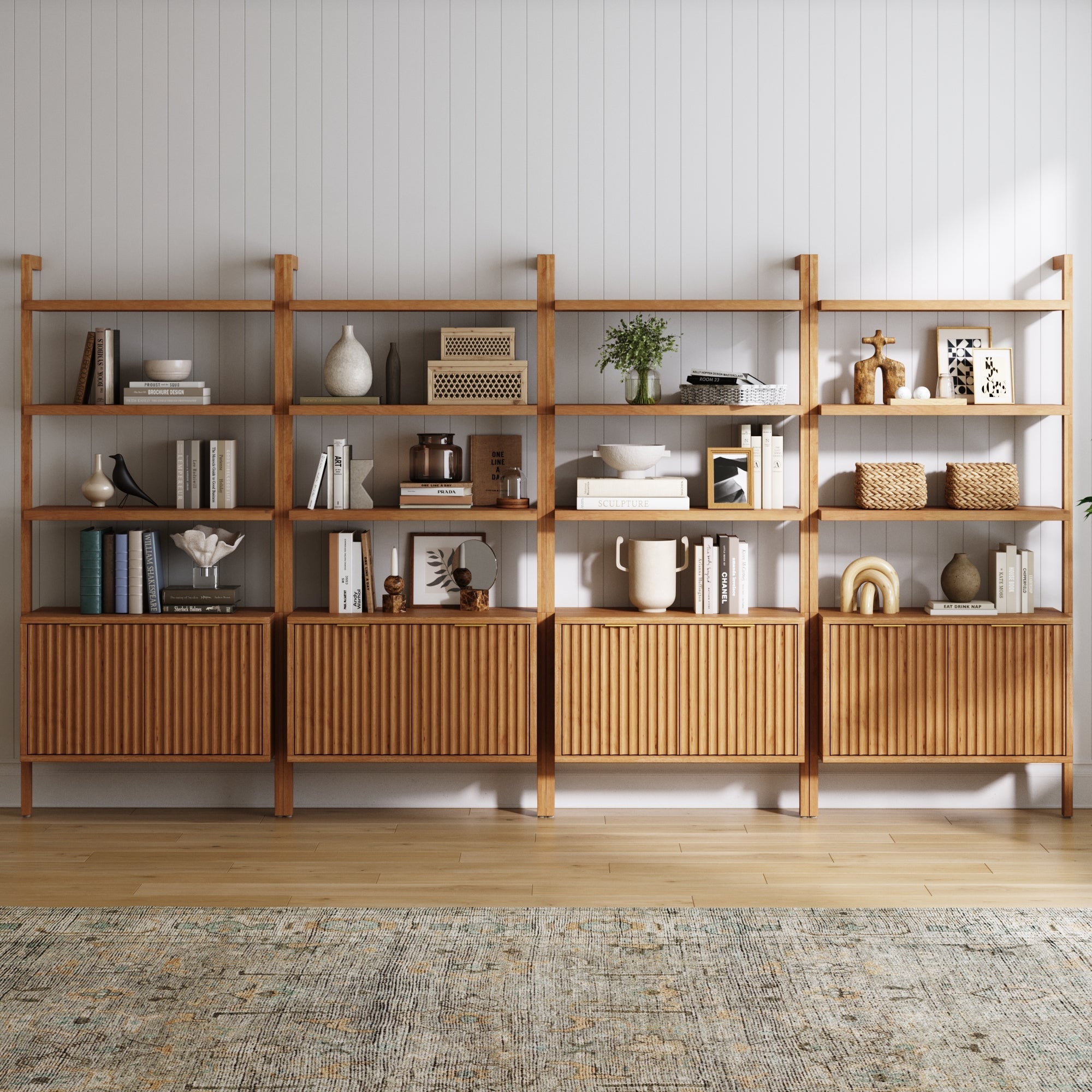 The Nathan James Mid-Century Fluted Wall Bookshelves with Doors, Warm Pine (Set of 4), display books, decor, plants, and vases against a white paneled wall, featuring storage cabinets below and set atop a patterned rug on a wooden floor.