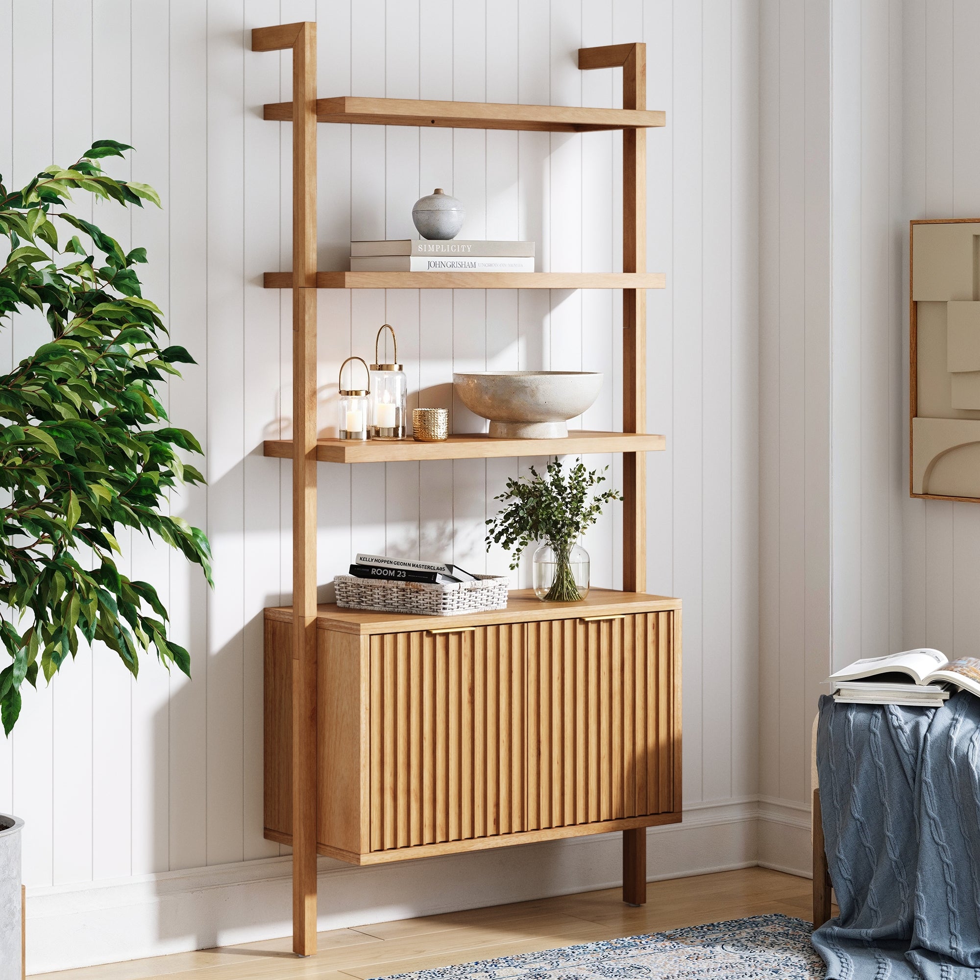 A modern wooden shelving unit with open shelves and a cabinet stands against a white paneled wall, displaying decorative items and plants. A leafy potted plant is nearby, adding greenery to the bright, cozy room.