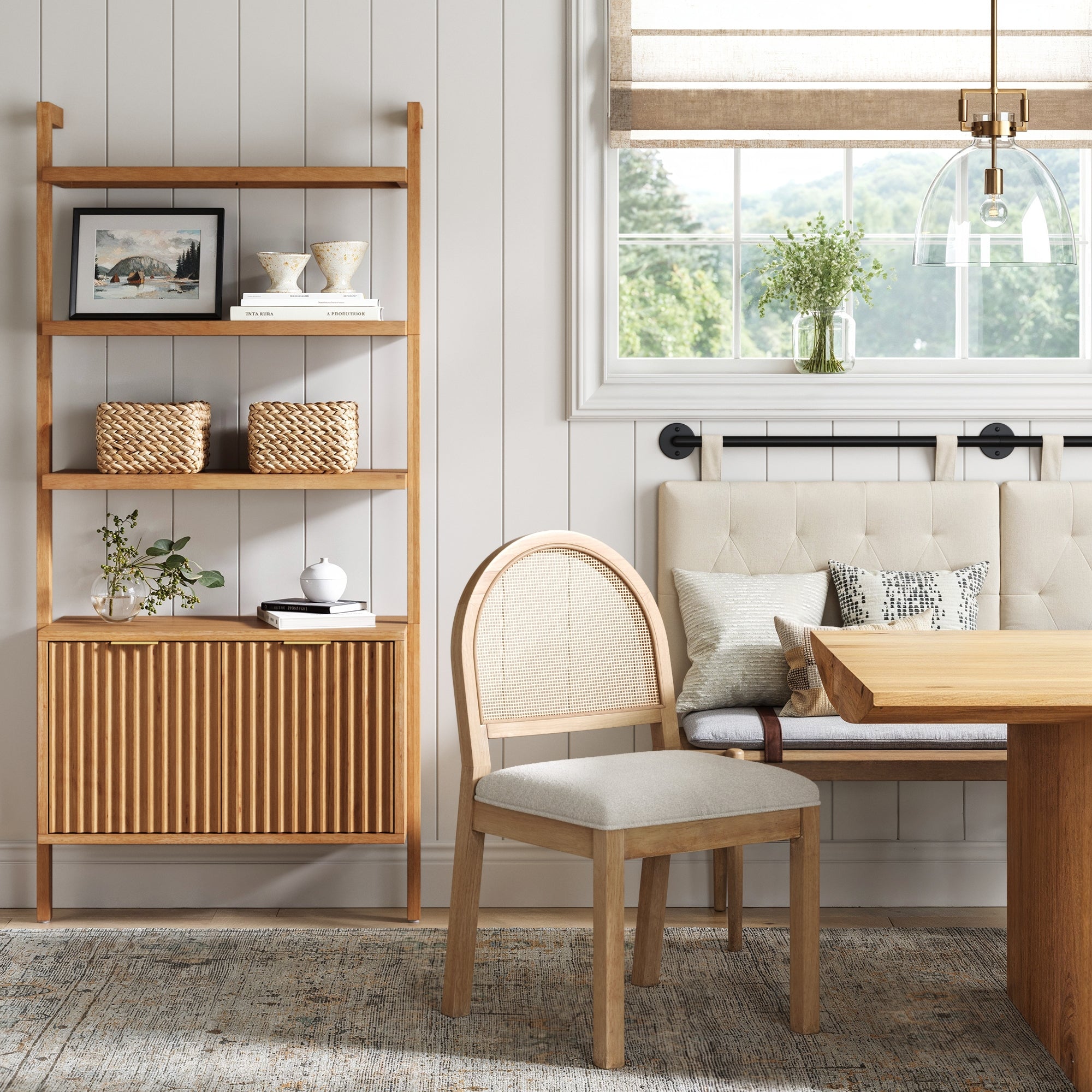 A cozy, modern dining nook with a wooden table, padded bench, neutral cushions, matching chair, and the Nathan James Mid-Century Fluted Wall Bookshelf with Doors in Warm Pine. Neutral colors and minimalist style enhanced by natural light.