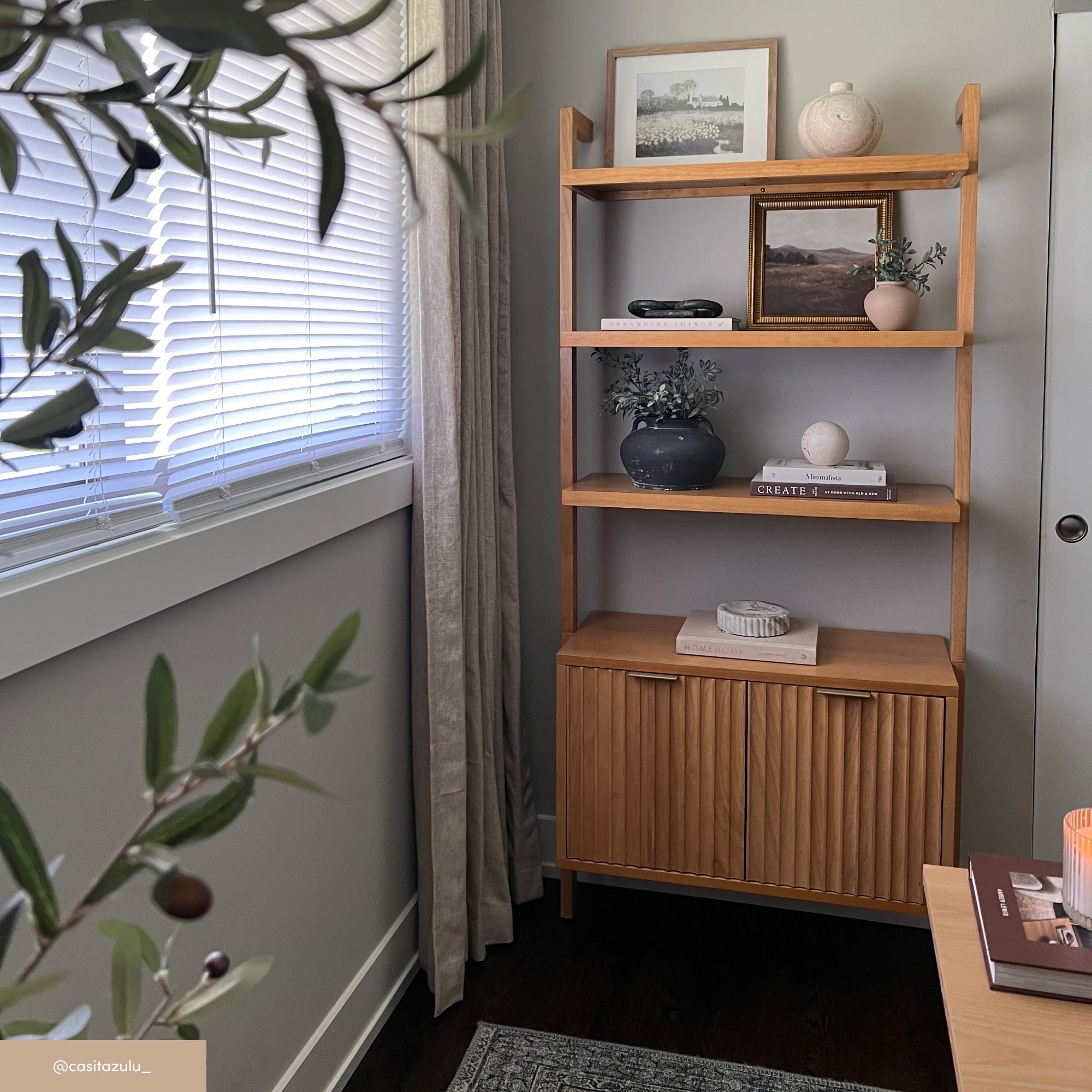 A wooden shelving unit in a cozy corner holds ceramics, framed art, books, and greenery. Sunlight filters through blinds, casting soft light on the neutral-toned, minimalist decor. An olive plant is in the foreground.
