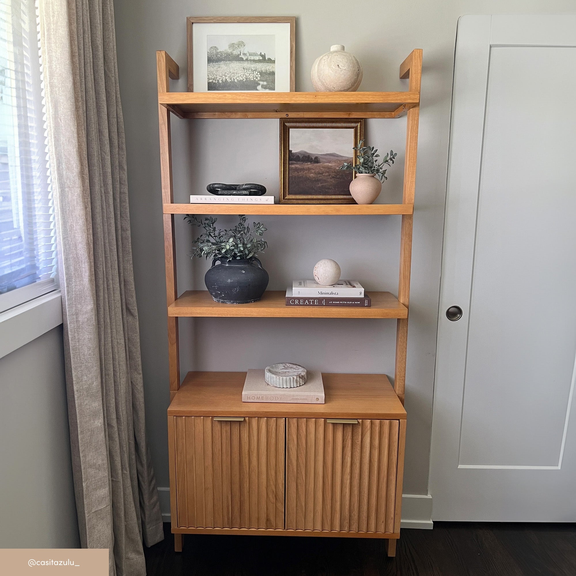 A light wood bookshelf with three shelves displays vases, framed photos, books, and greenery. Below is a cabinet with vertical grooves. The unit stands against a light wall near a window with beige curtains and a white door.