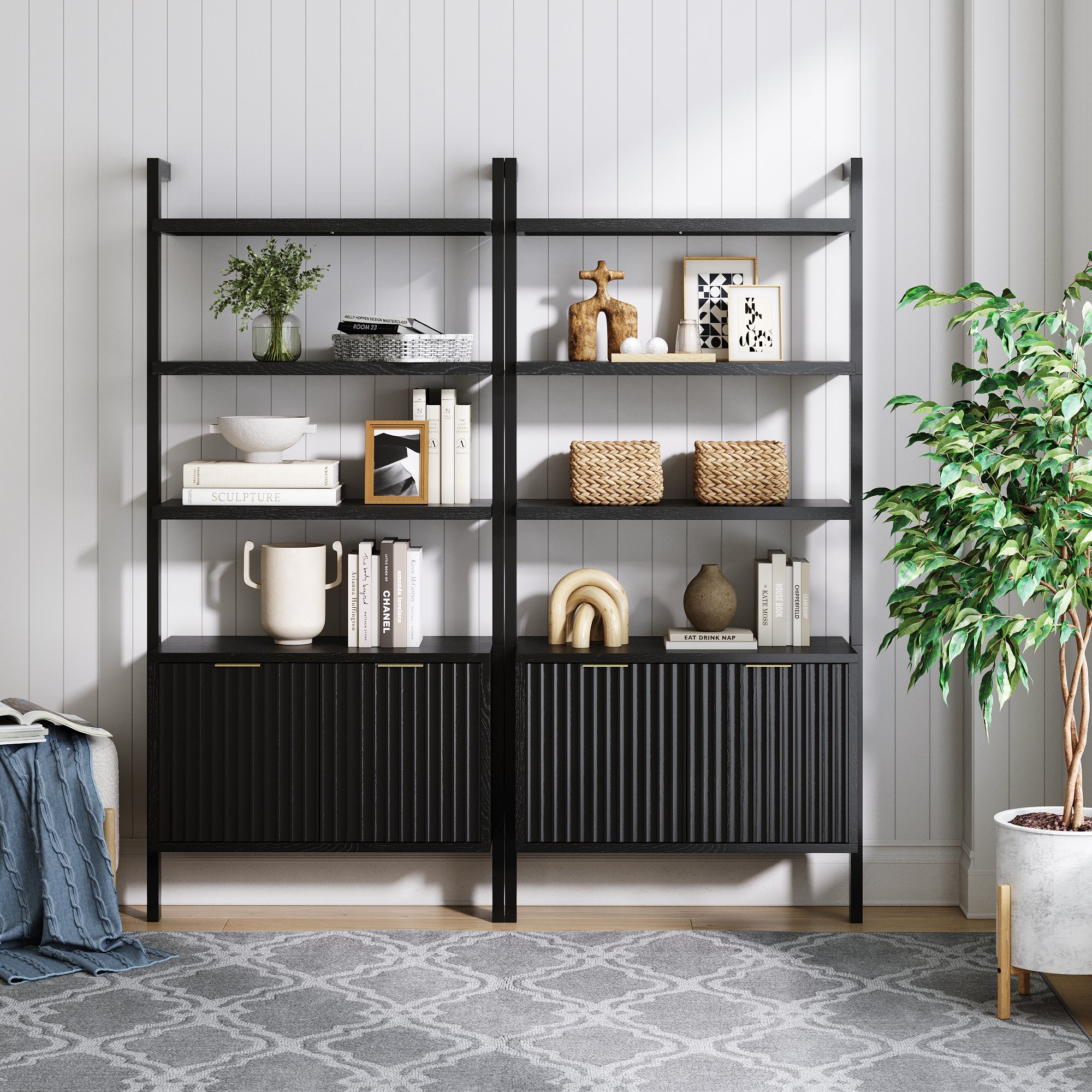 A modern black bookshelf with decor items, books, baskets, and vases stands against a white paneled wall. A leafy green plant sits to the right, and a gray patterned rug is on the floor.
