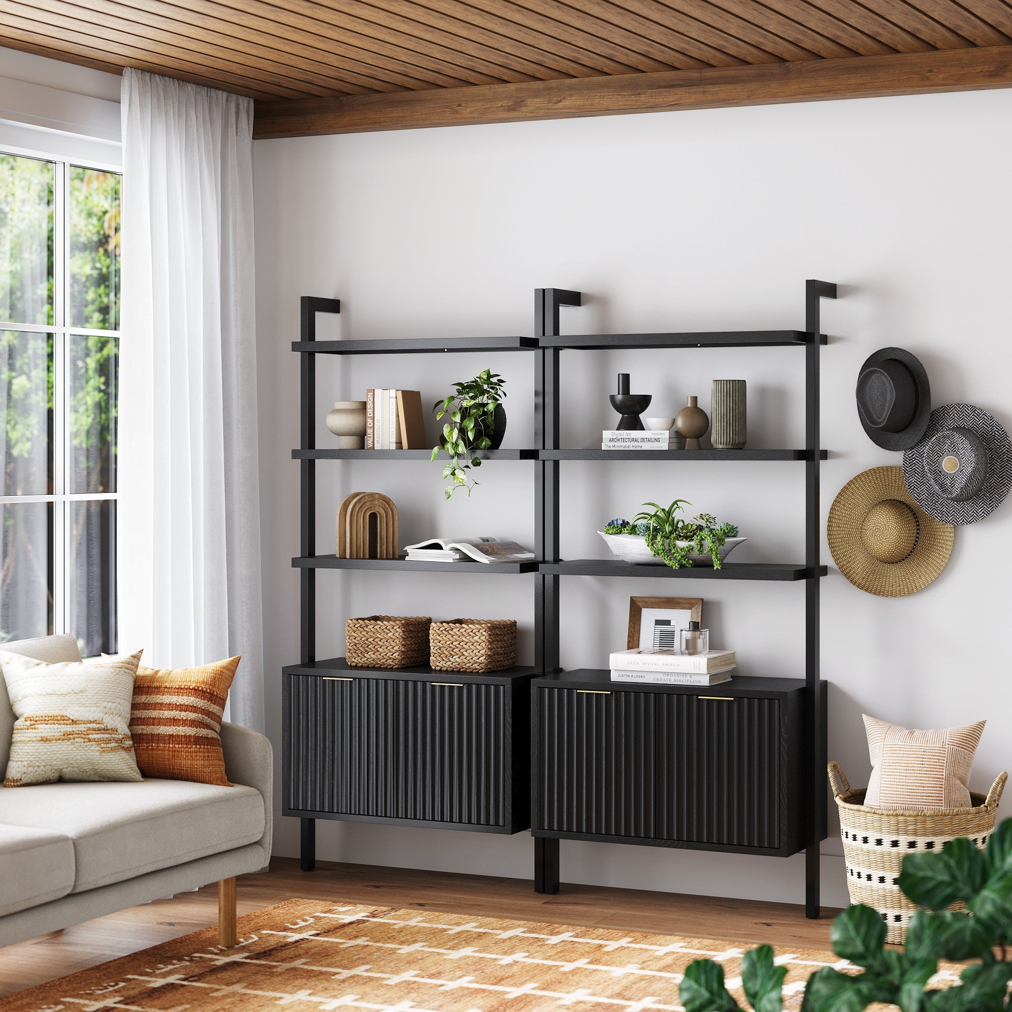 A modern living room showcases the Nathan James Mid-Century Fluted Wall Bookshelves with Doors Black (Set of 2) displaying plants, books, and decor. Two closed cabinets below offer storage. Nearby, hats hang and a neutral sofa sits on a textured rug by a large window.