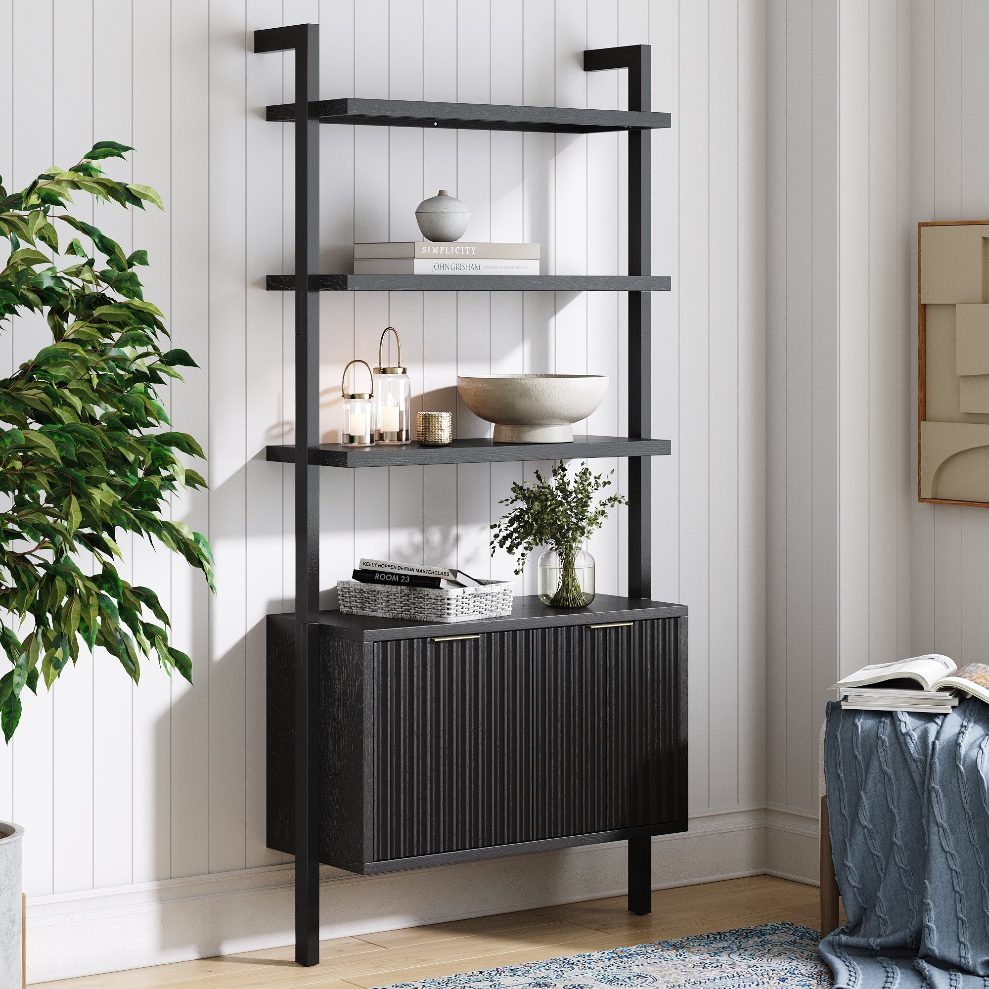 A modern black shelving unit with three open shelves and a closed cabinet stands against a white paneled wall. Decorative objects, books, and greenery are displayed on the shelves. A leafy plant sits nearby.