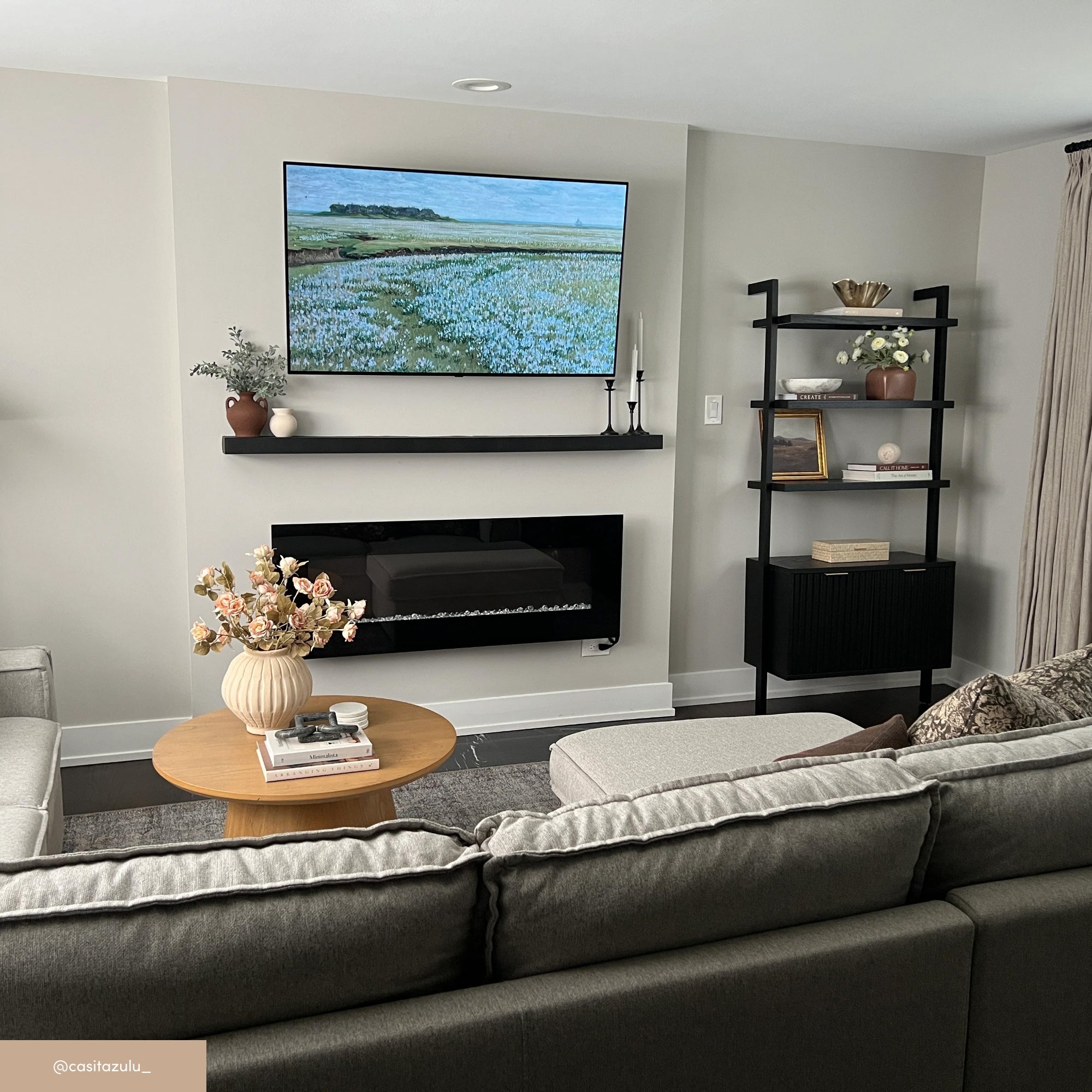 A modern living room with a gray sectional sofa, a round wooden coffee table with a floral arrangement, a wall-mounted TV displaying a landscape, black shelves with decor, and a black electric fireplace below the TV.