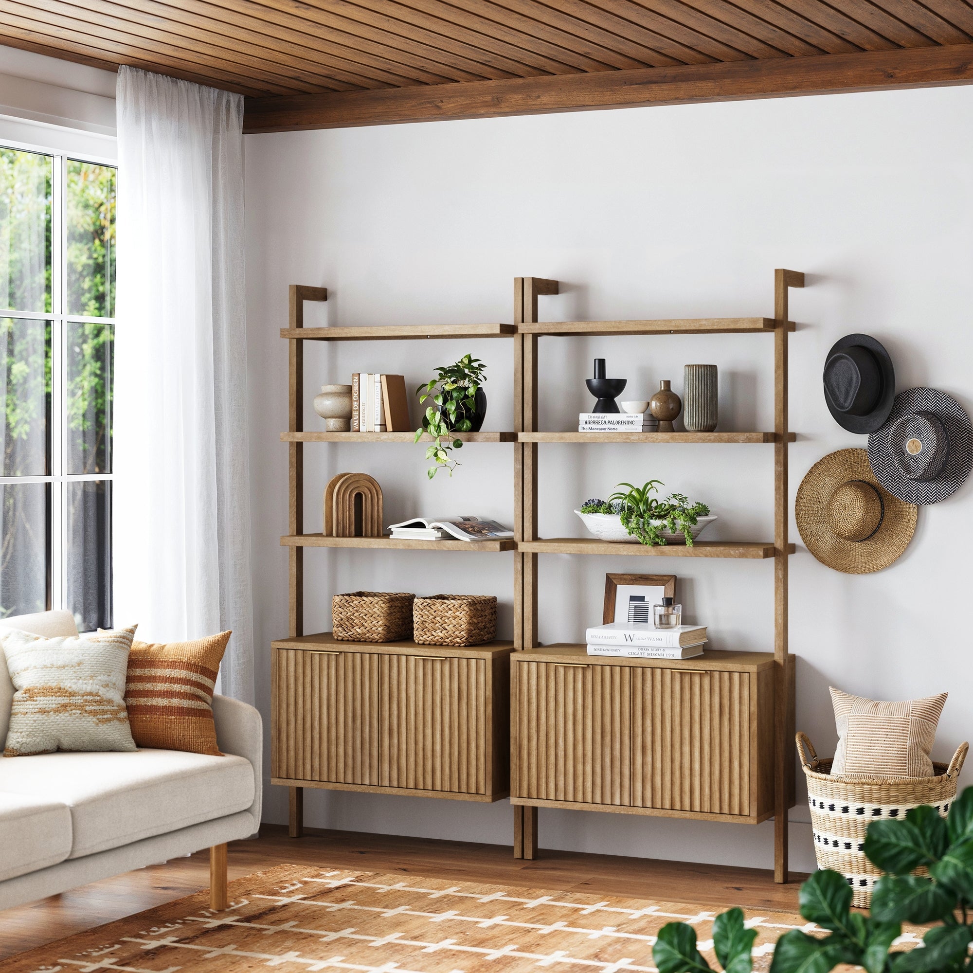 A modern living room features Nathan James Mid-Century Fluted Wall Bookshelves with Doors in light brown, displaying plants, books, and decor. Woven cabinets below, cozy sofa with pillows on the left, hats on the wall, and sunlight from large windows.
