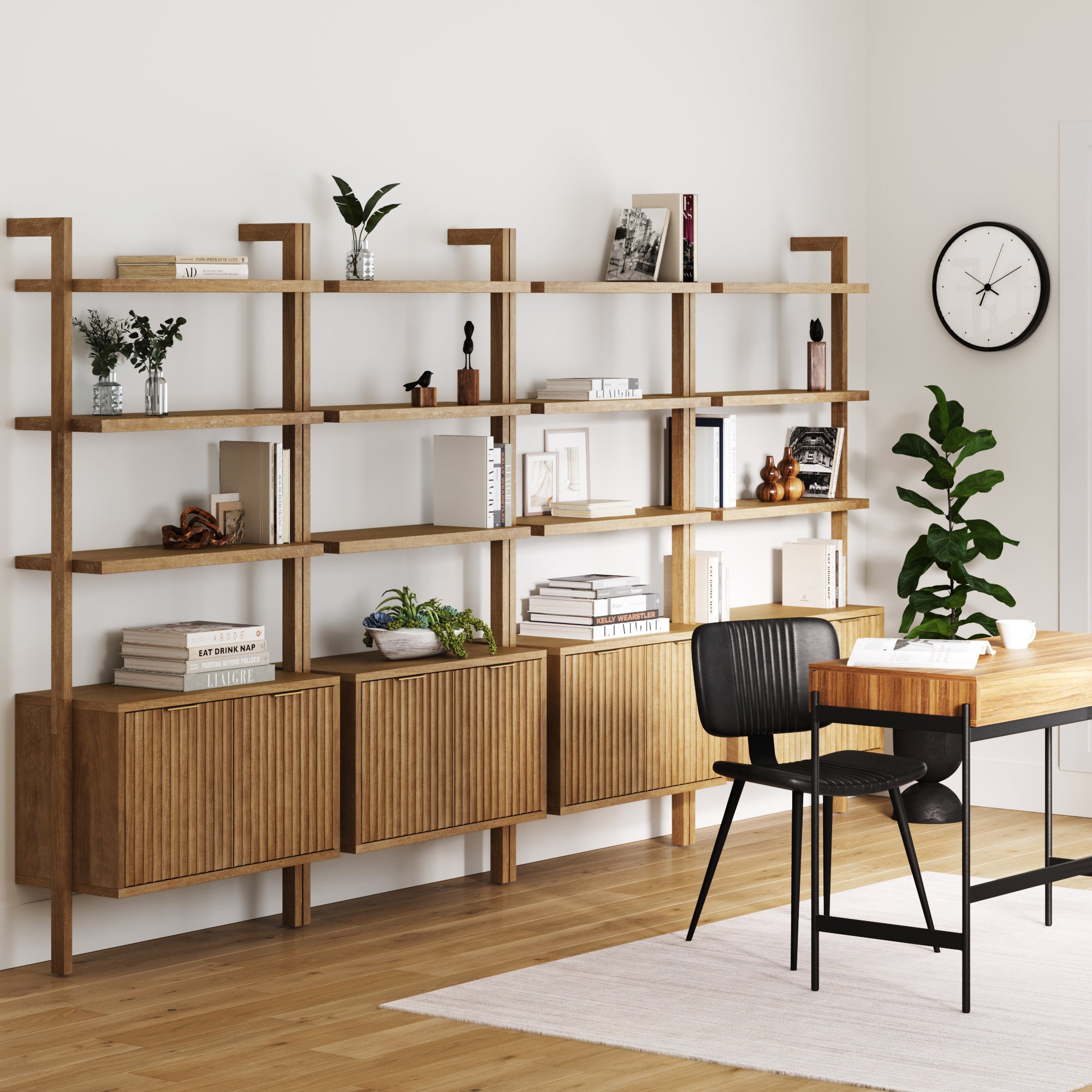 A modern home office features Nathan James Mid-Century Fluted Wall Bookshelves with Doors (Set of 4) in light brown, filled with books, decor, and plants next to a wooden desk, black chair, potted plant, and wall clock against a white wall.