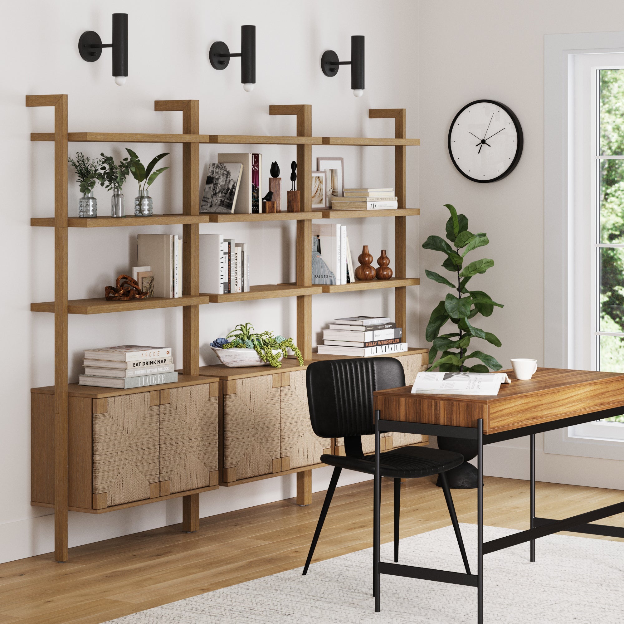 A modern home office features Nathan James Seagrass Wall Bookshelves with Doors in light brown (set of 3) holding books, plants, and decor, a wooden desk with a black chair, wall clock, large window for natural light, and three black wall lights above.
