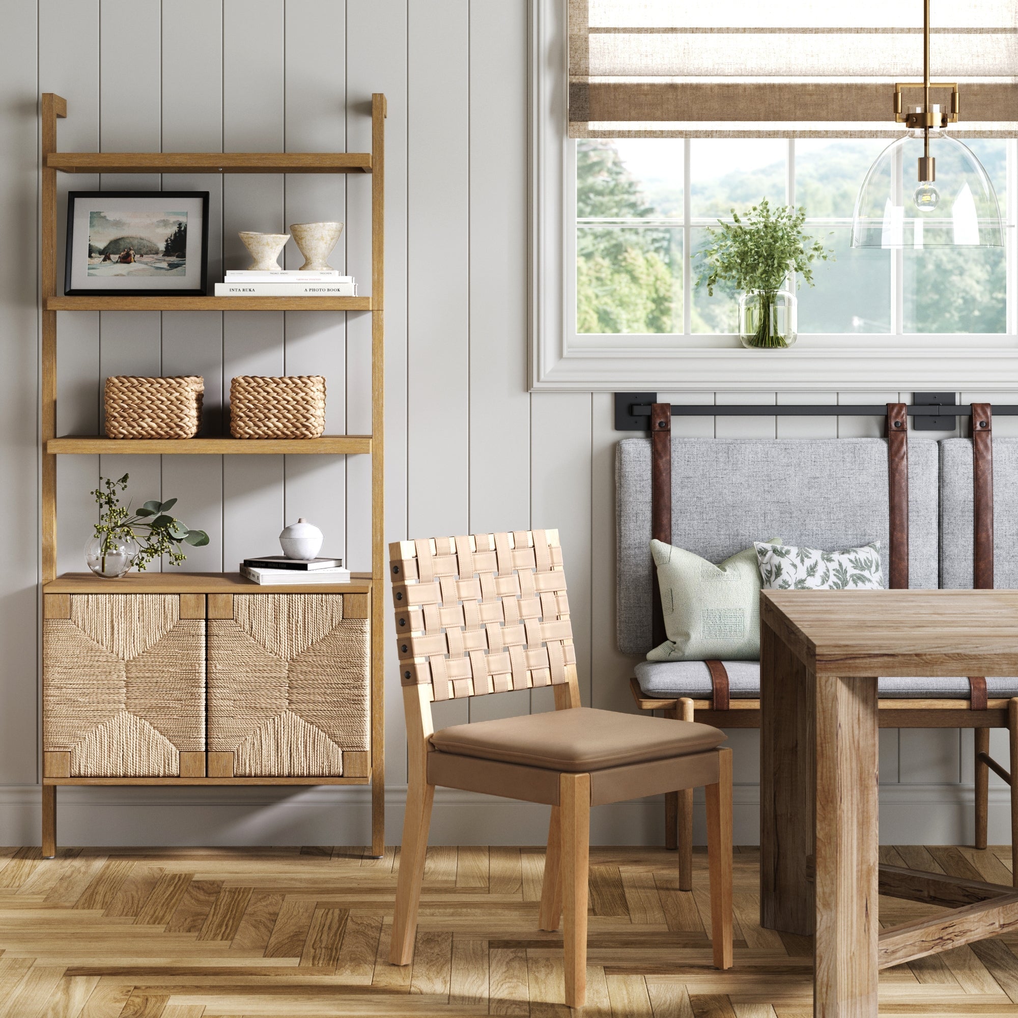 A modern dining area with a wooden table, woven chair, and bench. The Nathan James Seagrass Wall Bookshelf with Doors in Light Brown displays baskets, books, and art. A window shade filters light into the cozy, neutral space.