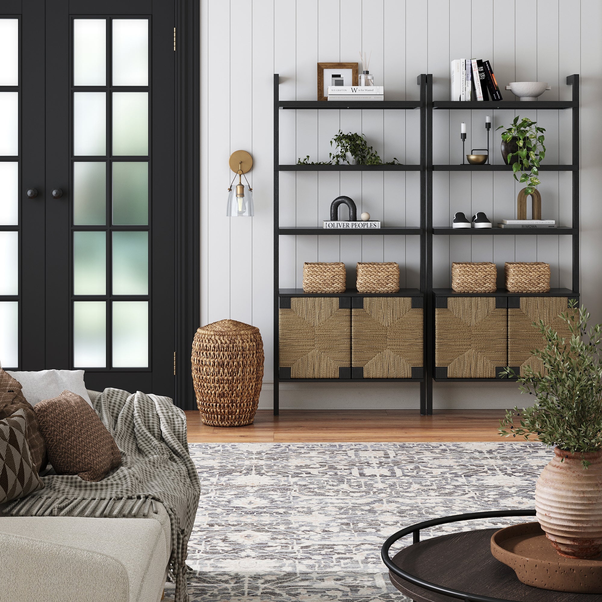 A modern living room features black Nathan James Seagrass Wall Bookshelves with Doors (Set of 2) styled with plants, books, and baskets, by a black French door and a sofa with patterned pillows and blankets on a light patterned rug.