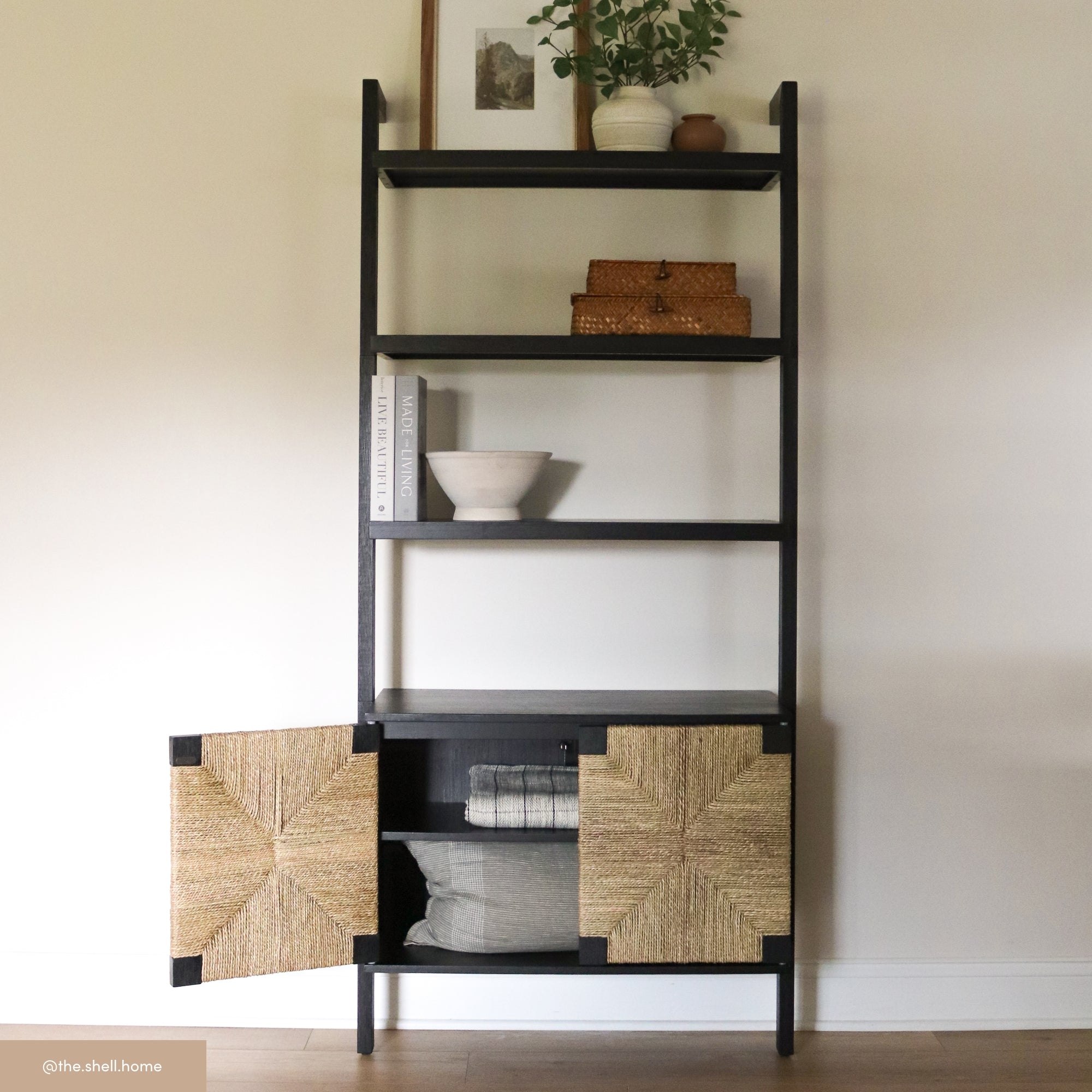 A black shelving unit with a plant, framed photo, woven baskets, books, and a ceramic bowl. The bottom section has two textured cabinet doors, one open to reveal pillows and folded linens inside.