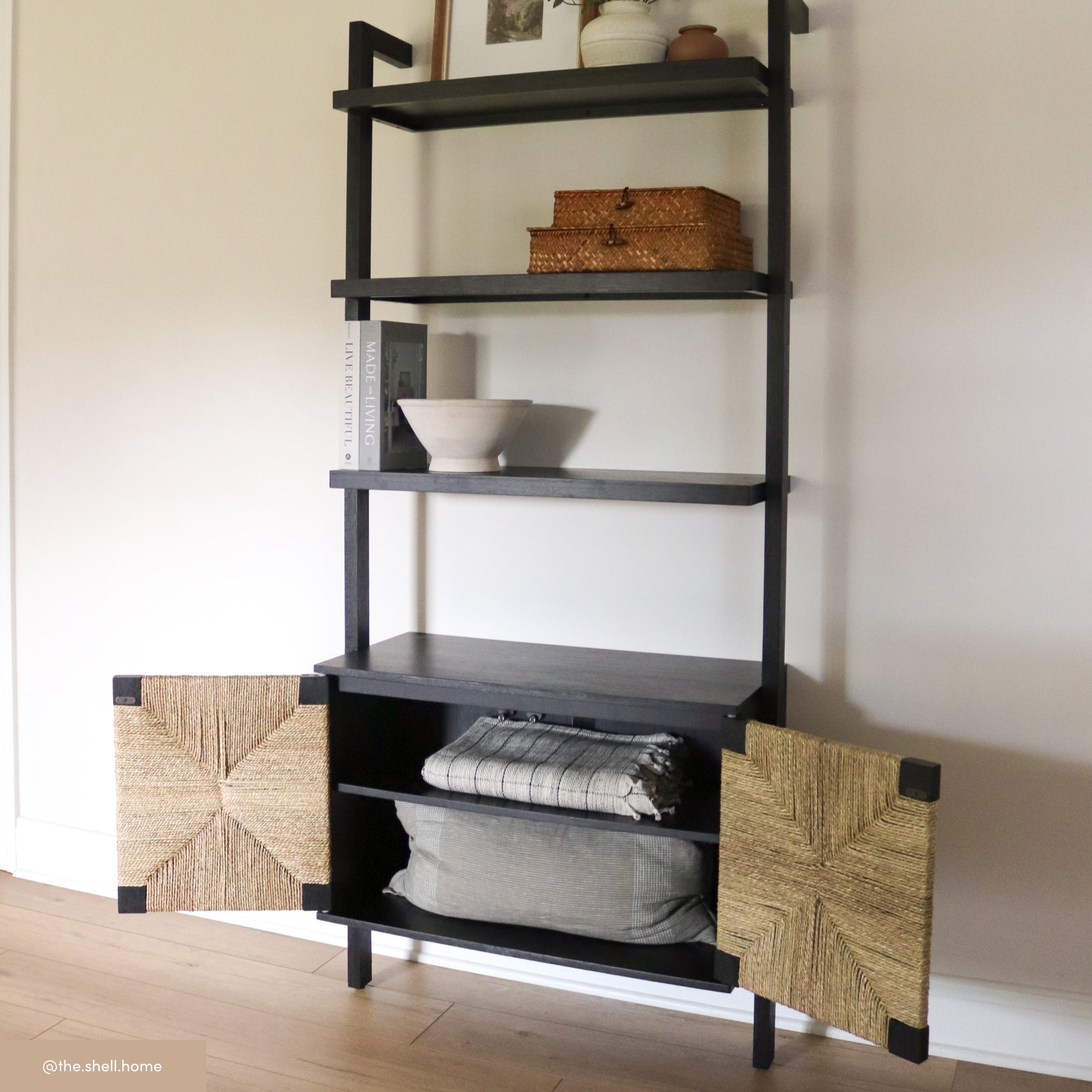 A black shelving unit with decorative items, including stacked baskets, a book, a bowl, and a vase on open shelves. The bottom cabinet has open doors showing folded blankets and a cushion inside.