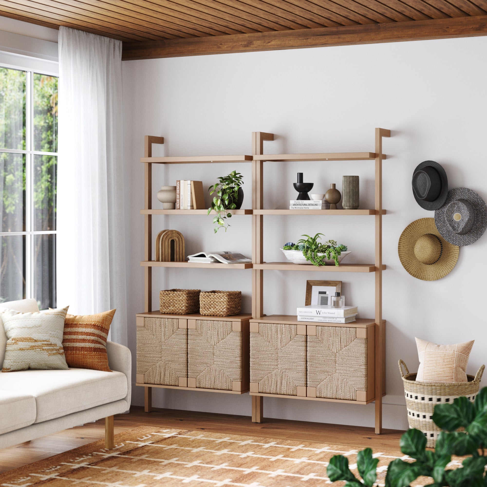 A modern living room features the Nathan James Seagrass Wall Bookshelf with Doors Raw Oak (Set of 2), displaying plants, baskets, books, and decor. A cream sofa with patterned pillows sits by a window, while three hats hang on the white wall.