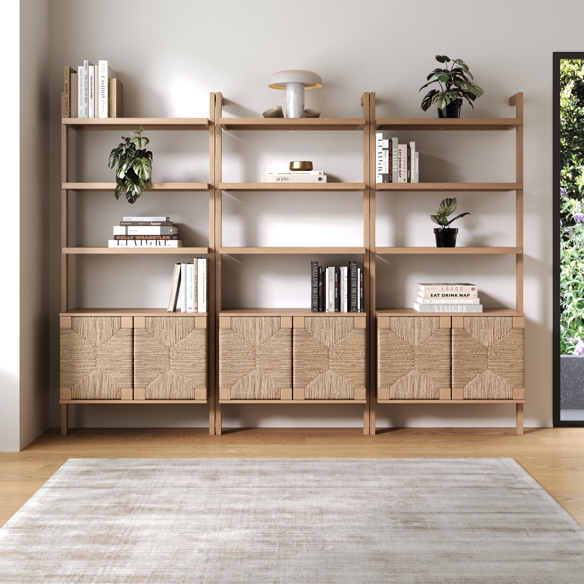 The Nathan James Seagrass Wall Bookshelf with Doors Raw Oak (Set of 3) showcases books, plants, and decor above four hand-woven seagrass cabinet doors, standing against a white wall on a light wood floor with a pale area rug.