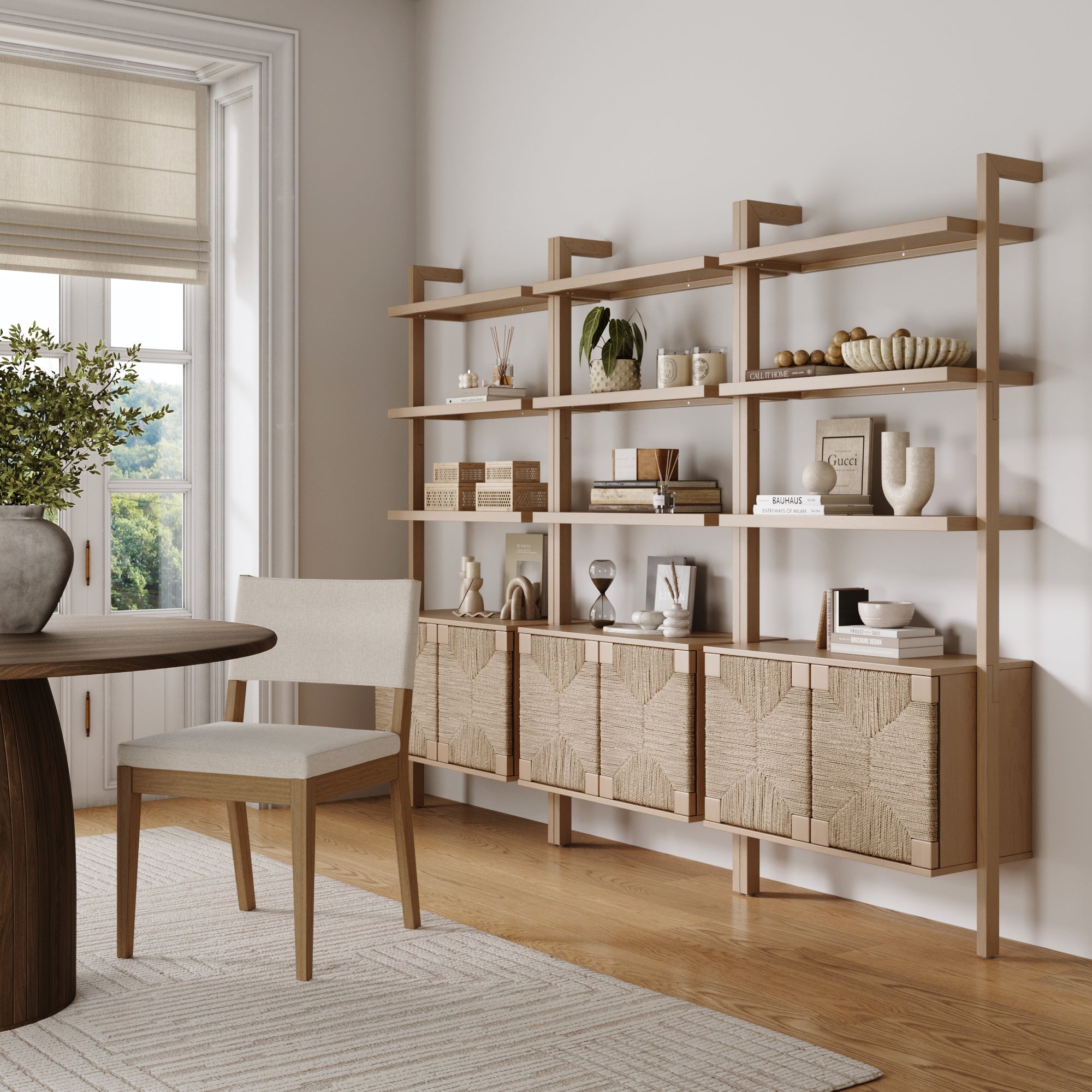 A modern, minimalist dining room features a raw oak table, light upholstered chair, and the Nathan James Seagrass Wall Bookshelf with Doors (Set of 3), styled with books and decor against a white wall and large window.