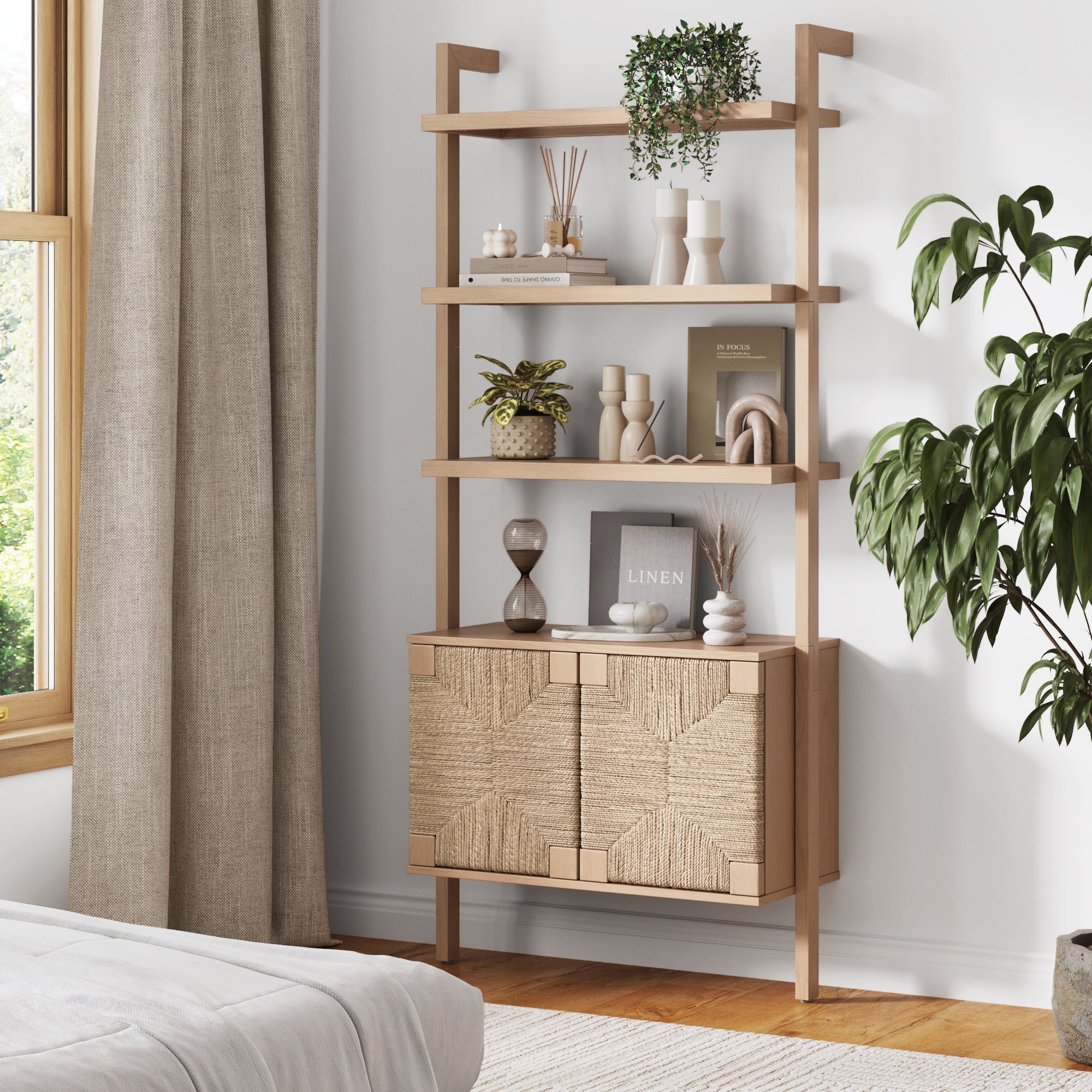 A Nathan James Seagrass Wall Bookshelf with Doors in raw oak displays plants, books, and decor against a white wall near a large leafy plant and beige curtains by the window. A bed with white bedding is partially visible in front.