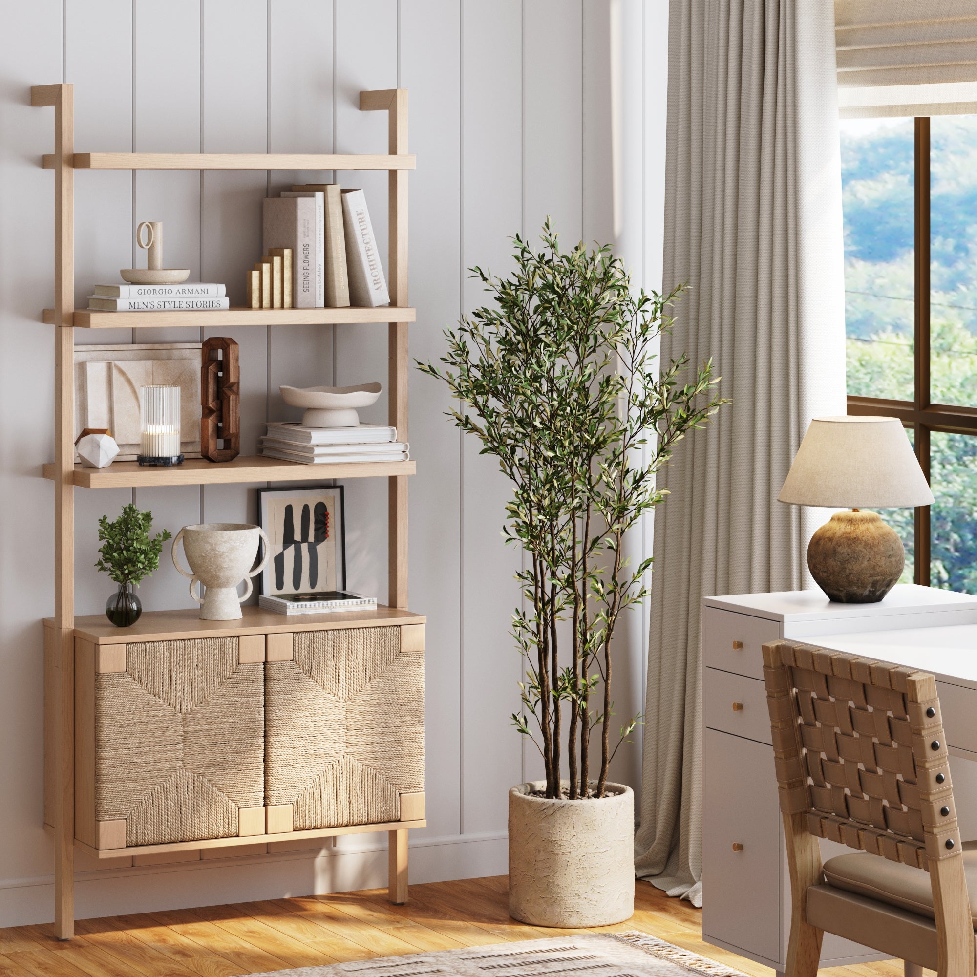 A cozy home office with a wooden desk, a woven chair, a potted plant, a lamp, and a wooden shelving unit holding books, art, and decor. Sunlight streams in through a window with beige curtains.