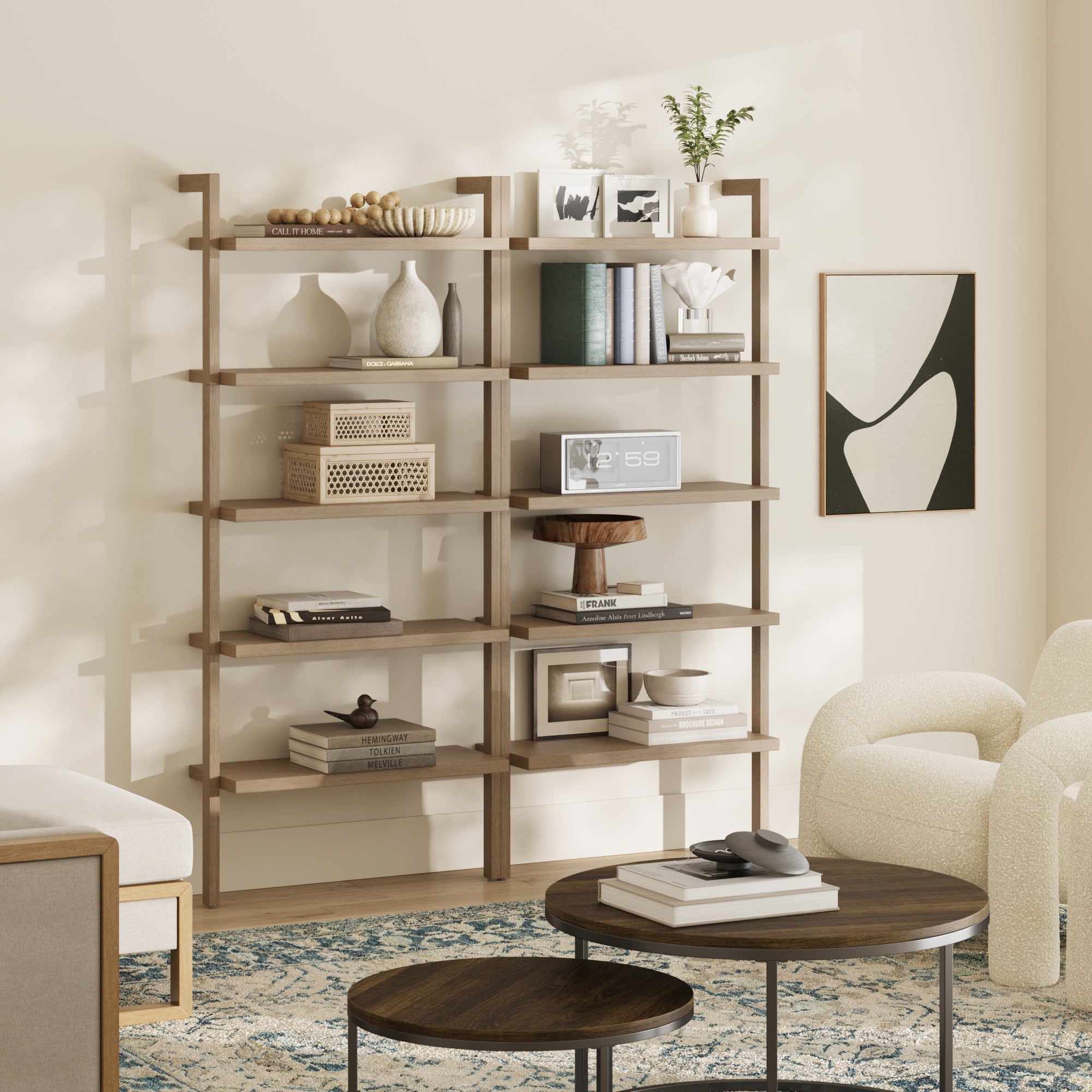 A modern living room features Nathan James Wood Wall Bookshelves in light brown with decor and books. A round coffee table with a tray and remote sits on a blue-patterned rug, surrounded by cream chairs and abstract wall art.