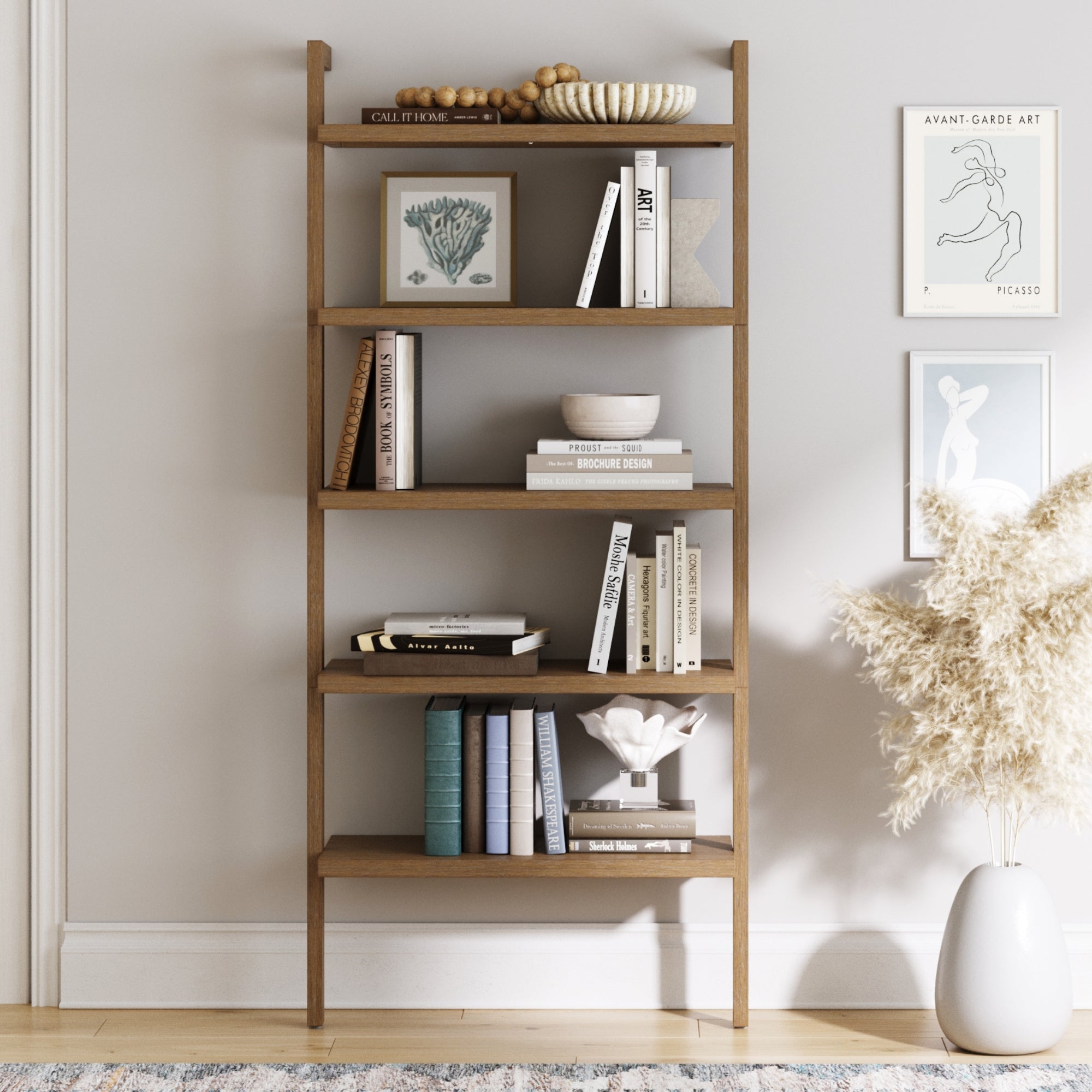 A wooden bookshelf against a light gray wall holds books, bowls, and decorative objects. Art prints and tall pampas grass in a vase are displayed nearby, creating a cozy, modern look.