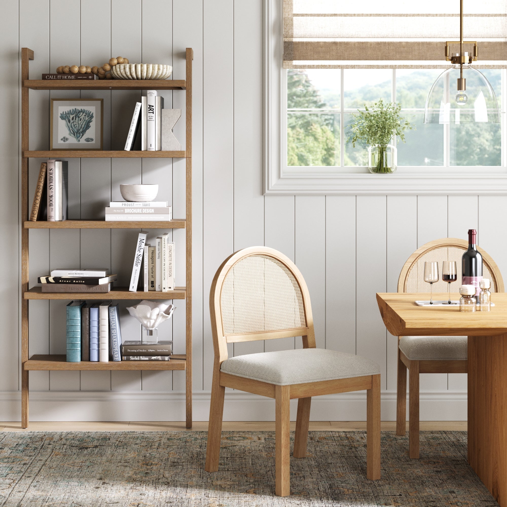 A cozy dining room with a wooden table, two chairs with wicker backs, a wall-mounted bookshelf with books and decor, a window with a beige shade, and wine set on the table. Light streams in through the window.