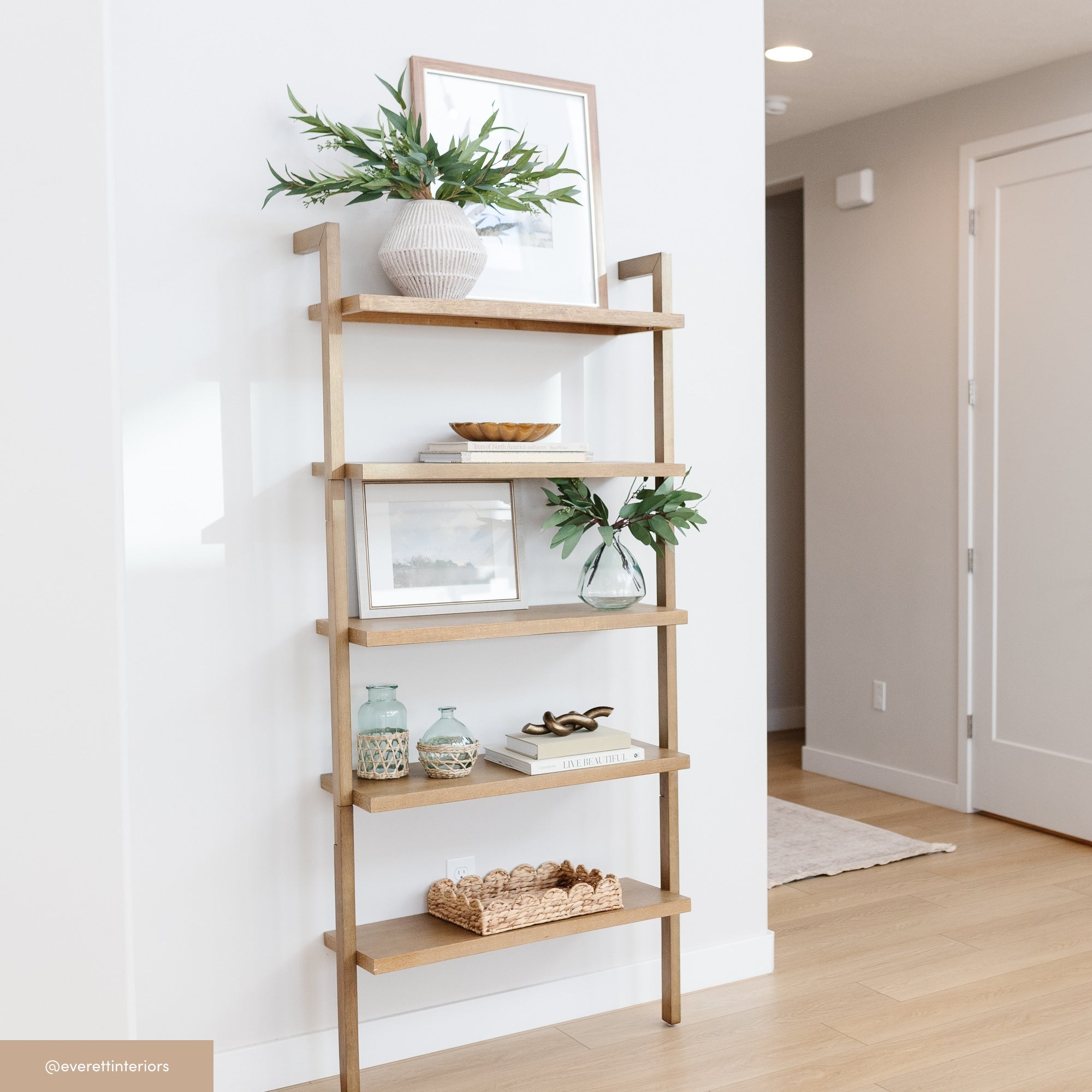 A wooden ladder-style shelf against a white wall holds framed art, potted plants, books, ceramic vases, a woven basket, and small decorative items in a bright, minimalist room with light wood flooring.