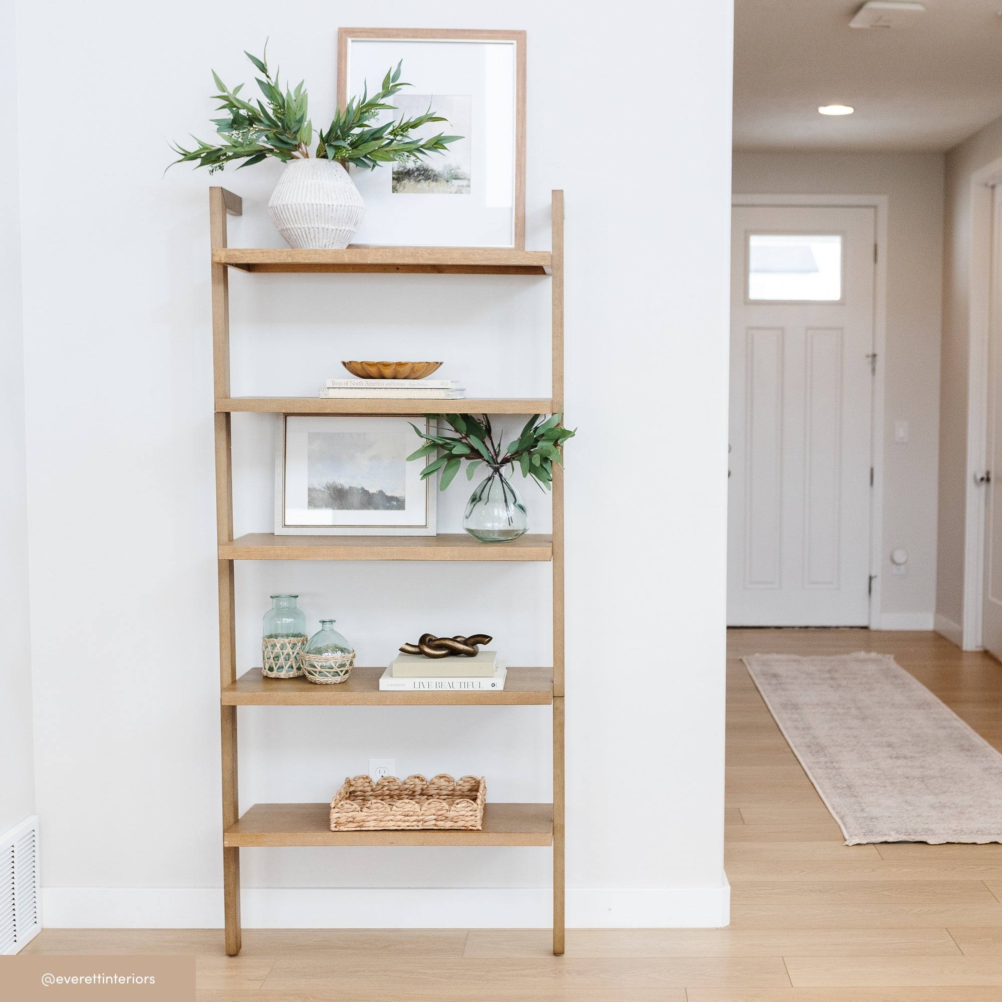 A wooden ladder-style shelf against a white wall holds decorative items including a vase with greenery, a framed picture, bowls, books, glass bottles, small baskets, and ornaments in a bright, modern entryway.