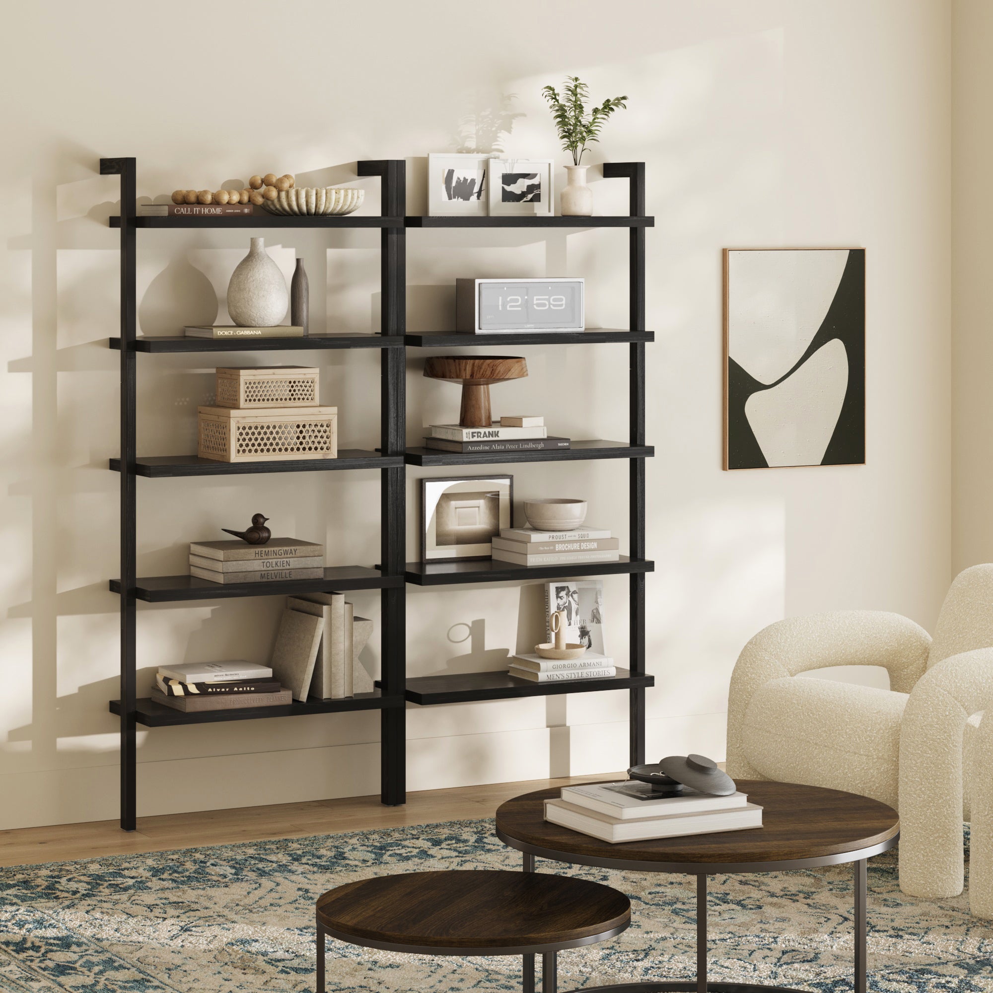 A modern living room features a Nathan James Wood 5 Tier Wall Mounted Bookshelf Black Oak (Set of 2) displaying books, décor, and plants. Two round wooden coffee tables and a cream textured armchair sit on a patterned rug with abstract art on the wall.