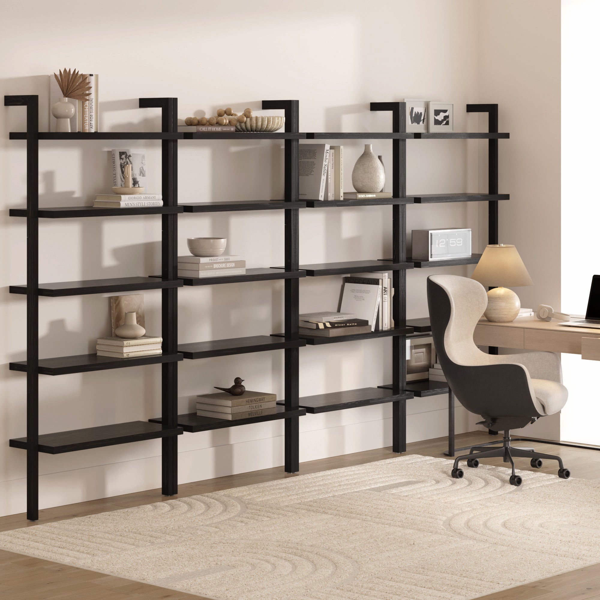 A bright, minimalist home office features a Nathan James Wood 5 Tier Wall Mounted Bookshelf (Set of 4) in black oak holding books and decor, with a desk, chair, lamp, and clock nearby on a light rug.