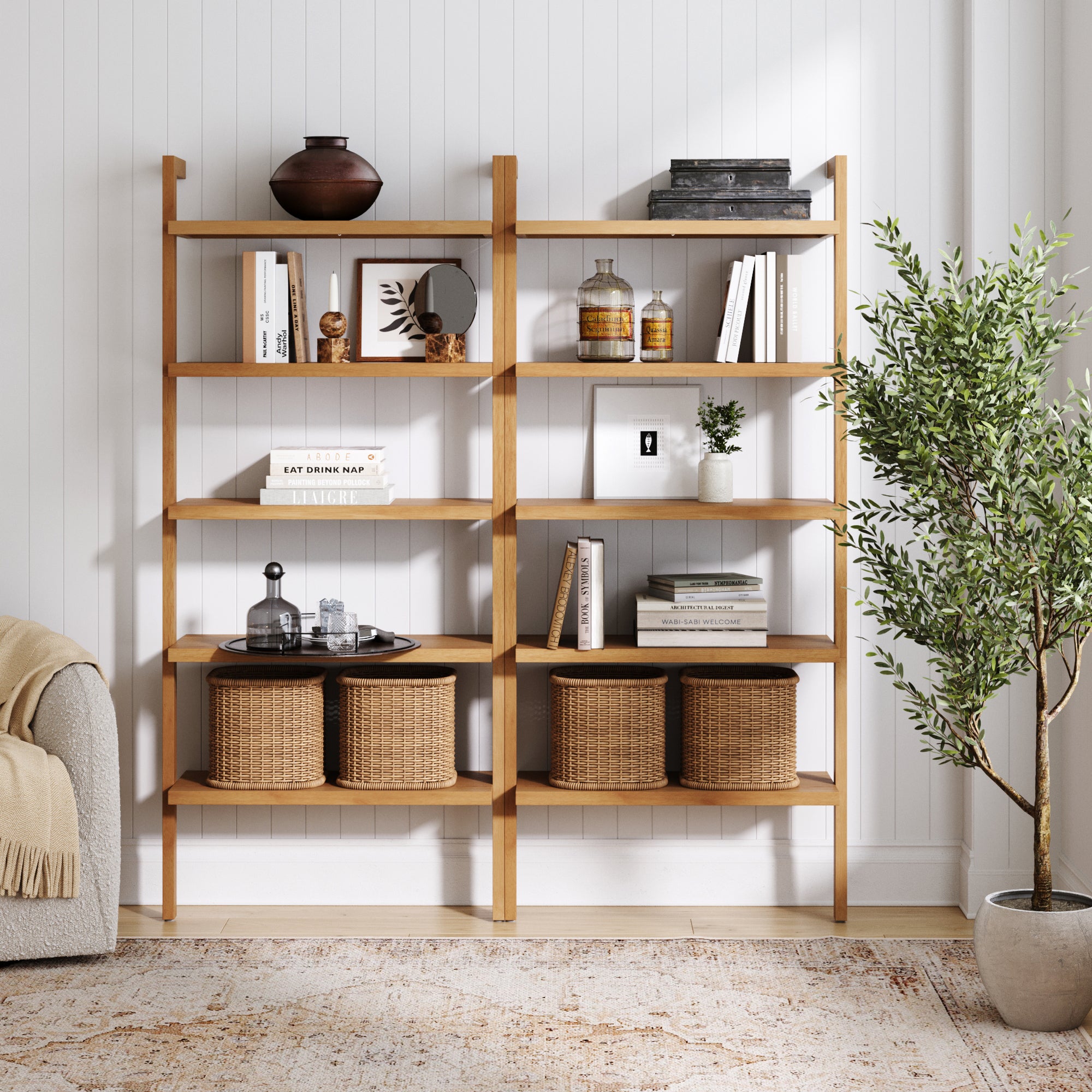 A pair of Nathan James Wood 5-Shelf Ladder Wall Bookshelves in warm pine hold books, decor, baskets, and vases against a white paneled wall beside a leafy plant and light armchair on a textured rug.