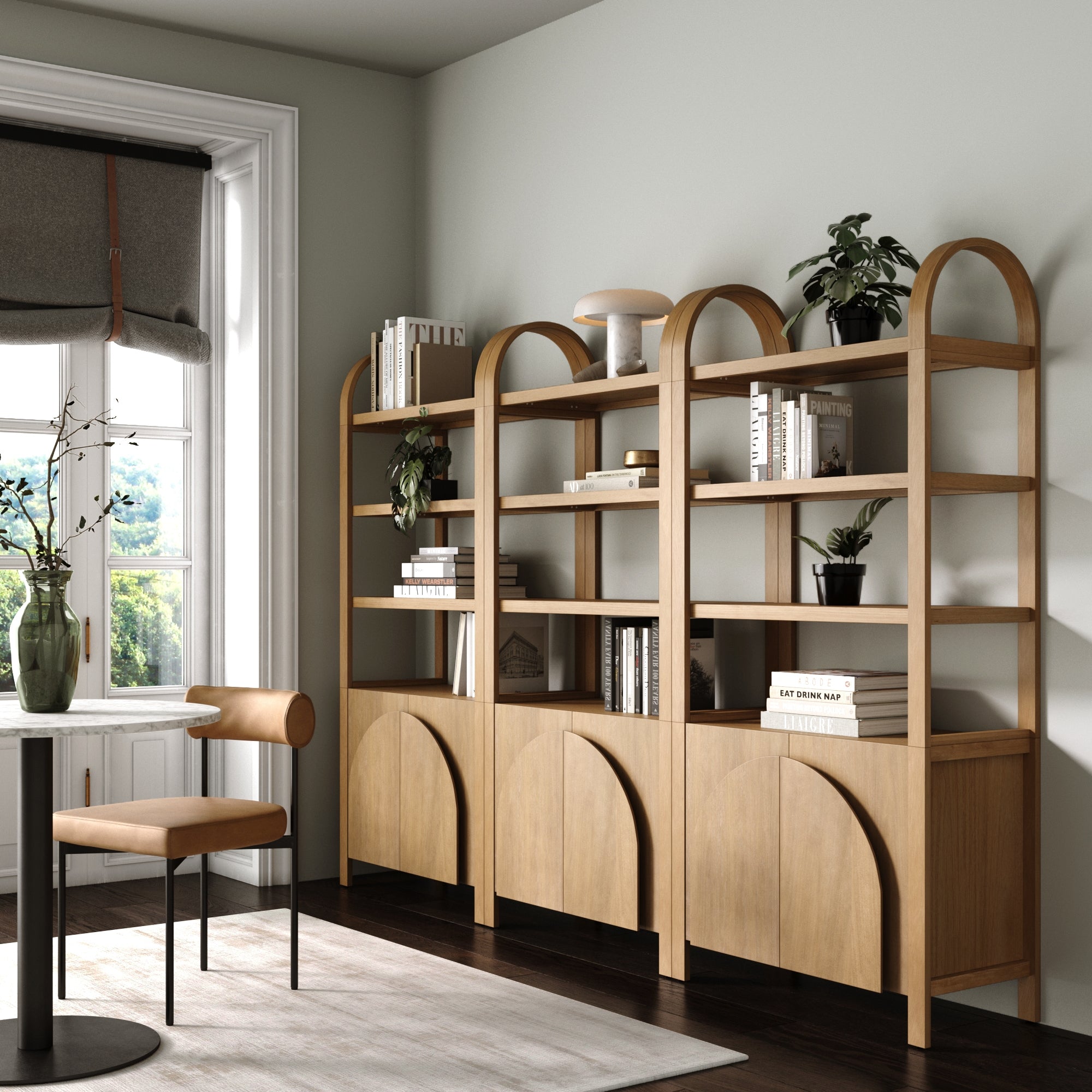 A modern, minimalistic study room featuring a set of three Nathan James Mid-Century Arched Bookshelves with Doors in light brown, open shelves with books and plants, a tan chair, small desk, and large window for natural light.
