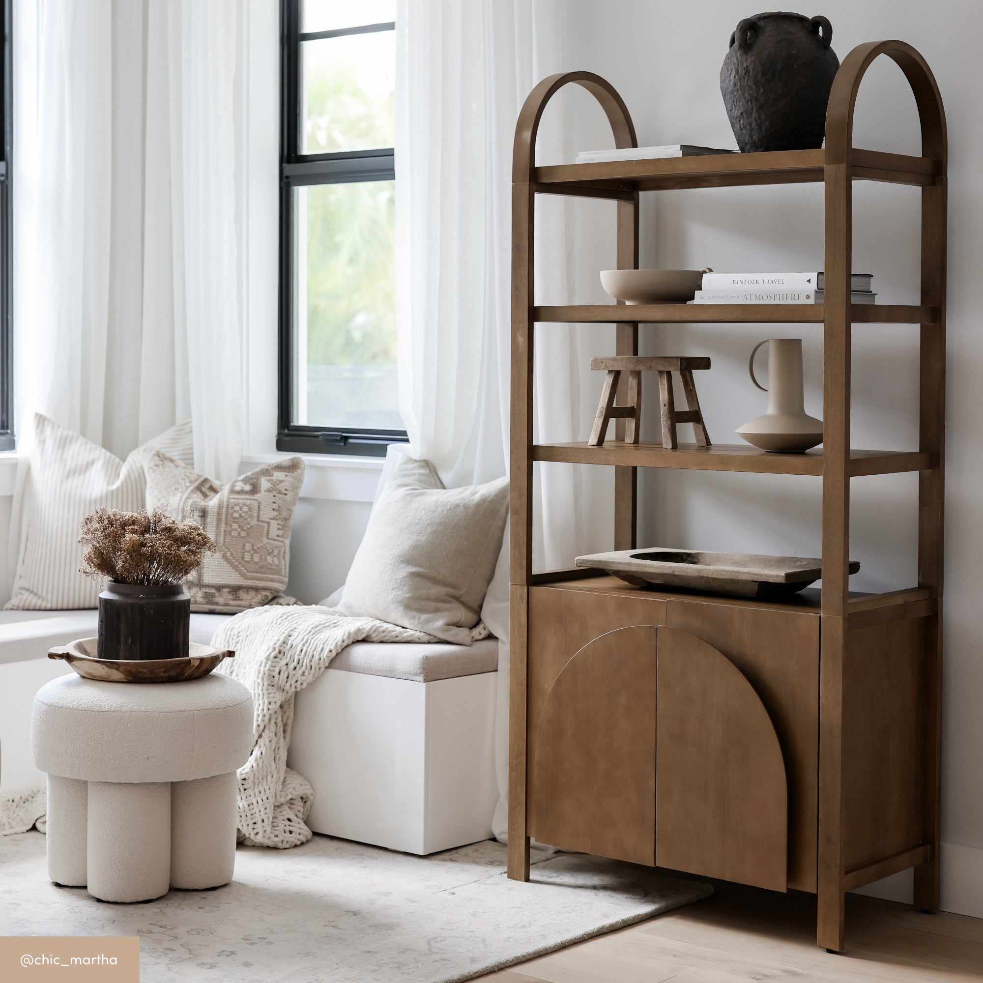 A cozy living space with a wooden shelving unit holding pottery and decor, a cushioned bench with neutral pillows and a knit throw, and a round textured stool topped with dried flowers, all in soft, natural tones.
