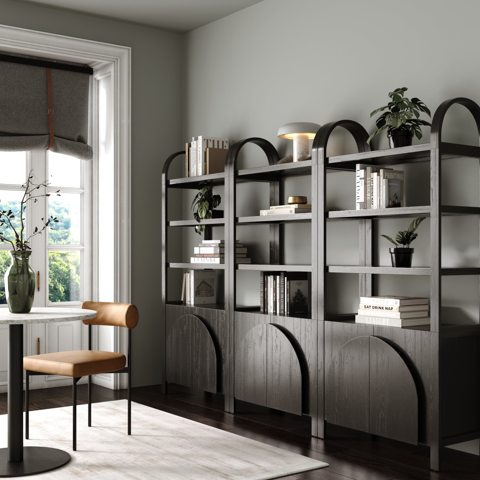 A modern room showcases three Nathan James Mid-Century Arched Bookshelves with Doors in black, organized books, plants, and a lamp. A brown chair by a round table sits near a window with a gray shade that lets in natural light.